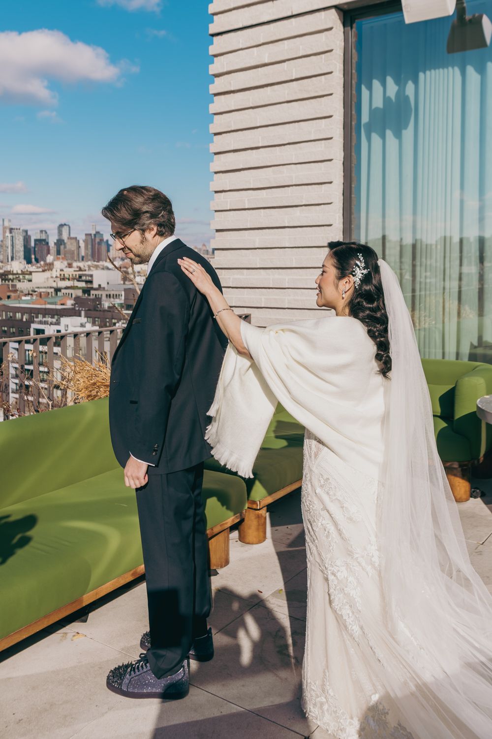 A bride in a white lace gown and cape surprises her groom on a rooftop with city skyline views.