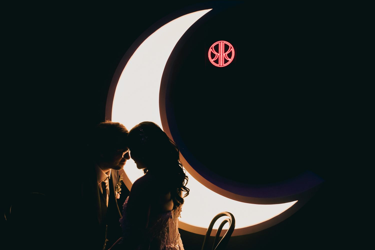 Bride and groom silhouette kissing in front of illuminated crescent moon backdrop.
