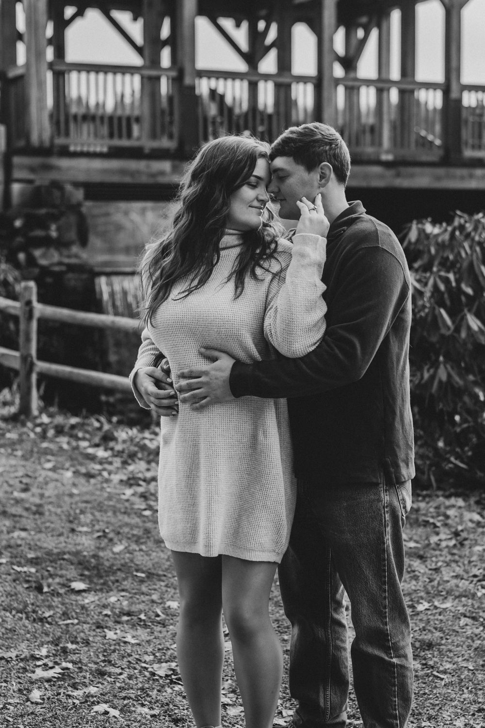 Black and white photo of a romantic couple embracing outdoors near a rustic wooden structure.