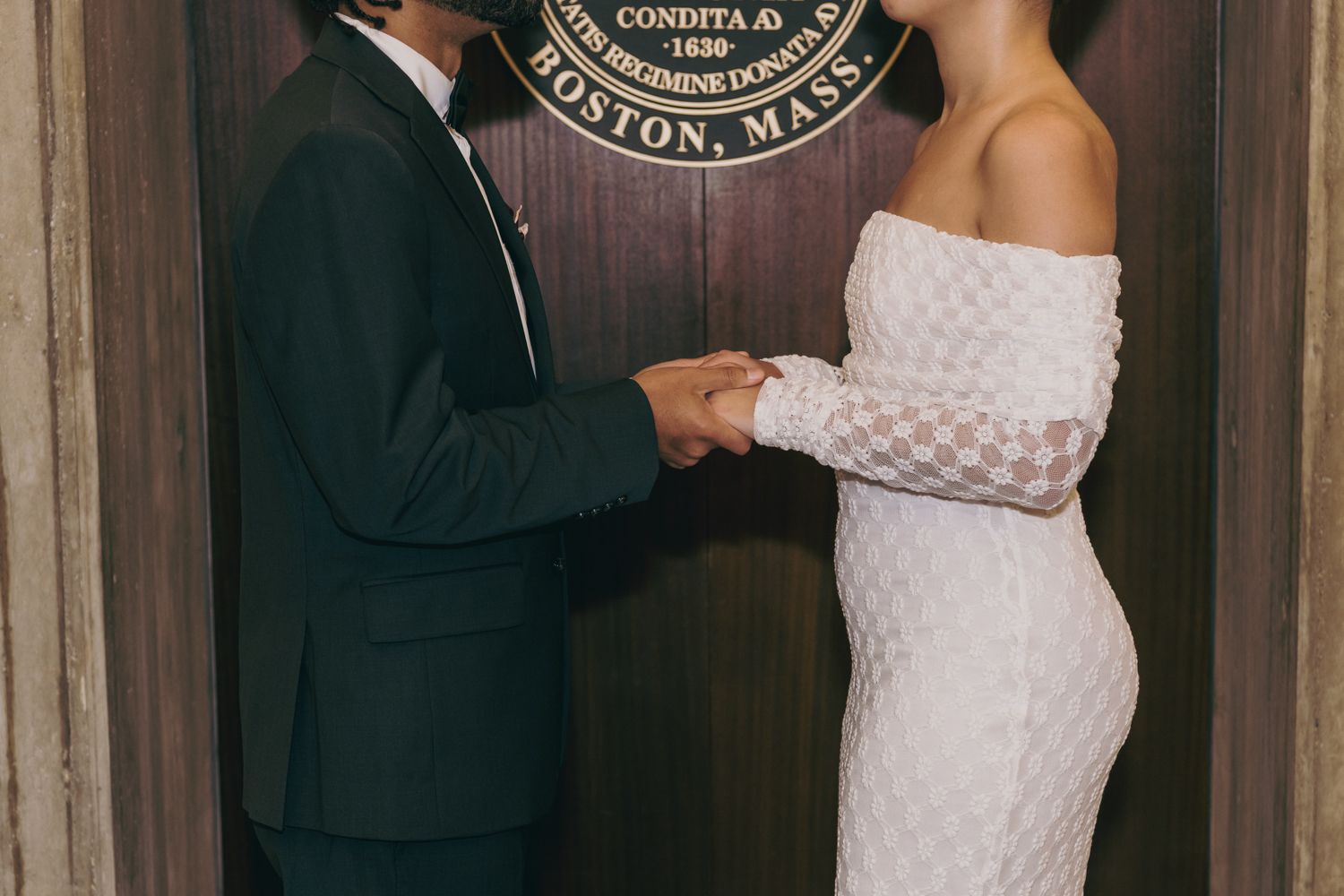 Couple holding hands in wedding attire before a Boston, Massachusetts seal on a wooden door.