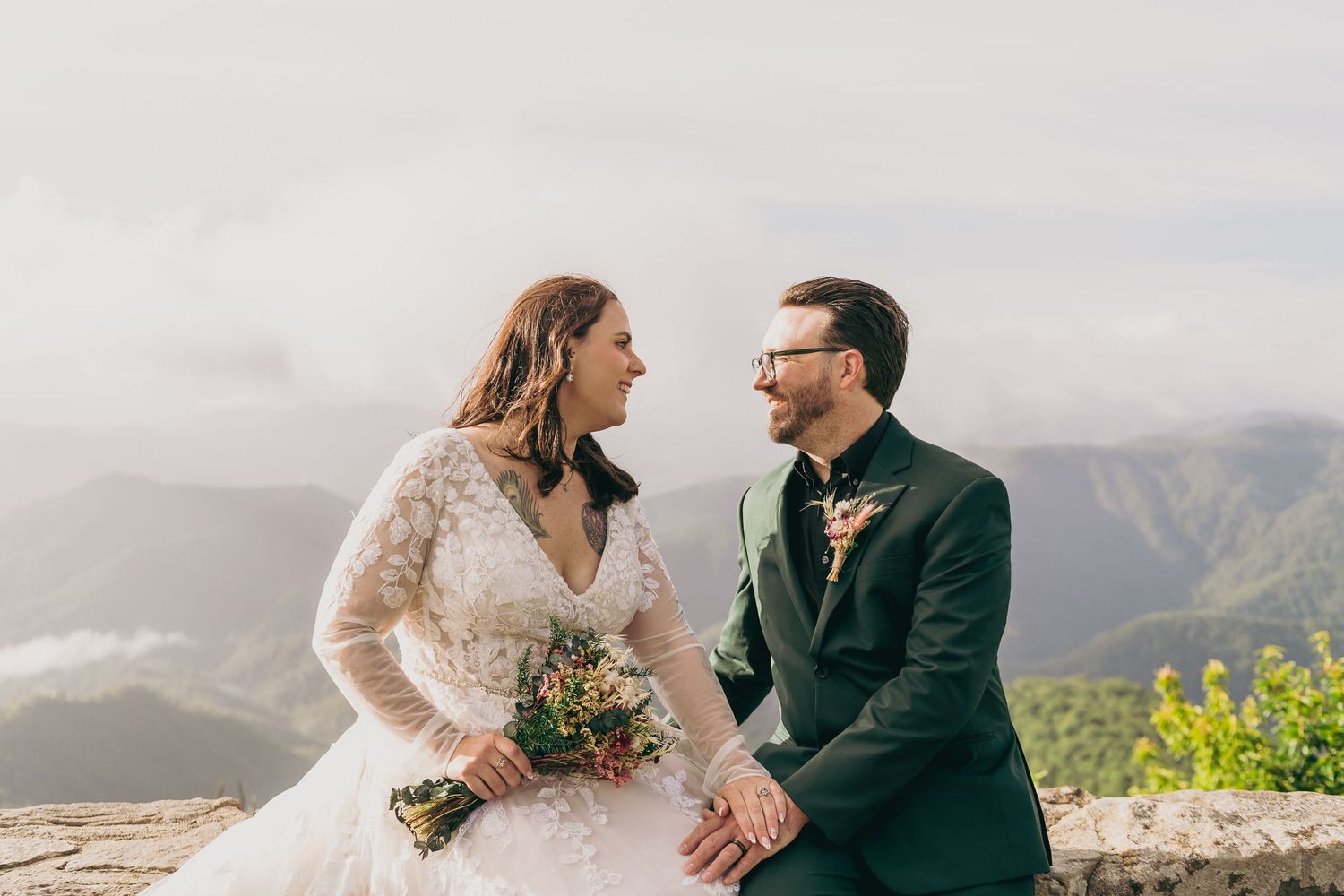 Bride and groom share a joyful moment on a mountain overlook, with lush green valleys and cloudy skies behind them.
