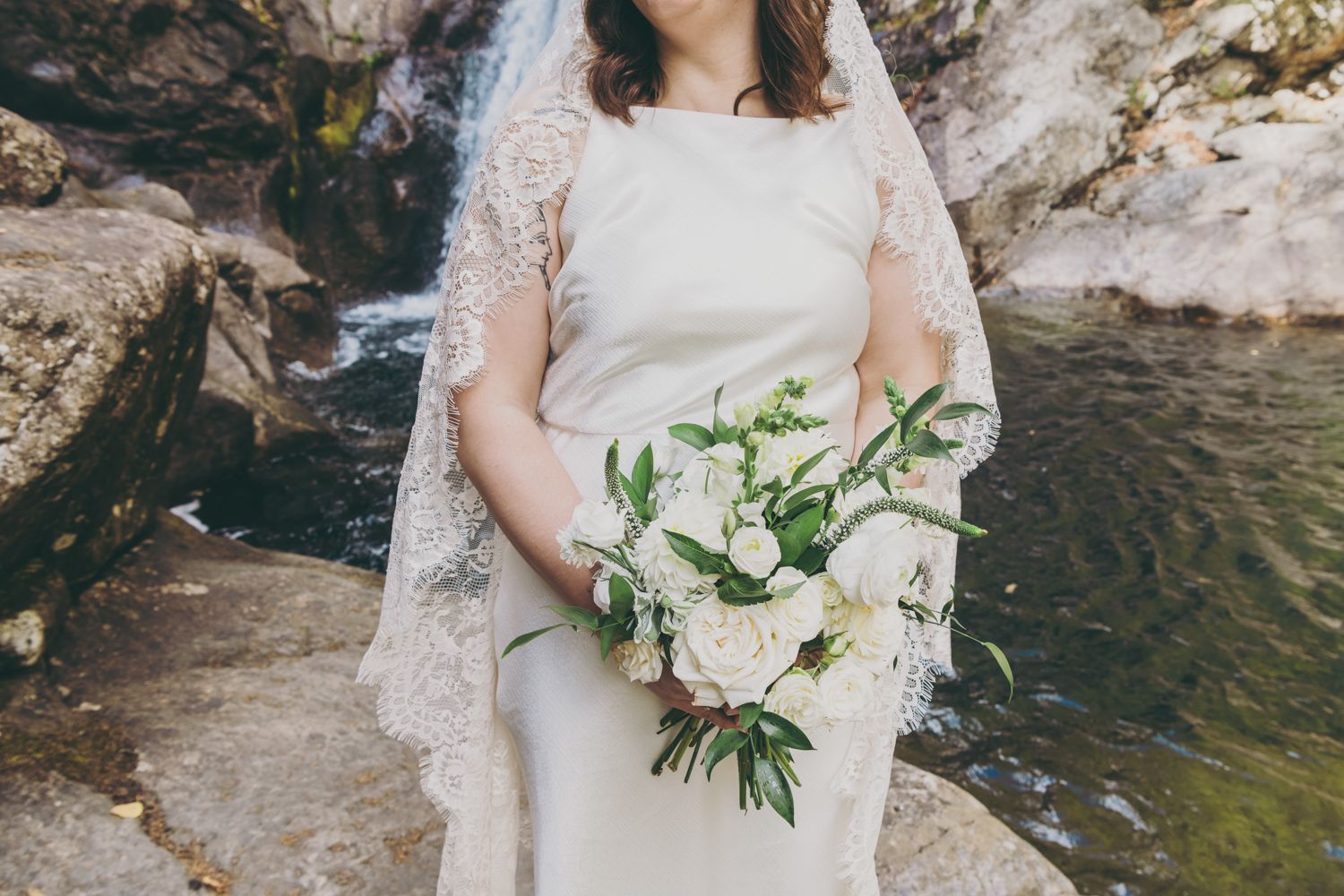 Bride in white gown and lace veil holds elegant white bouquet near a waterfall.