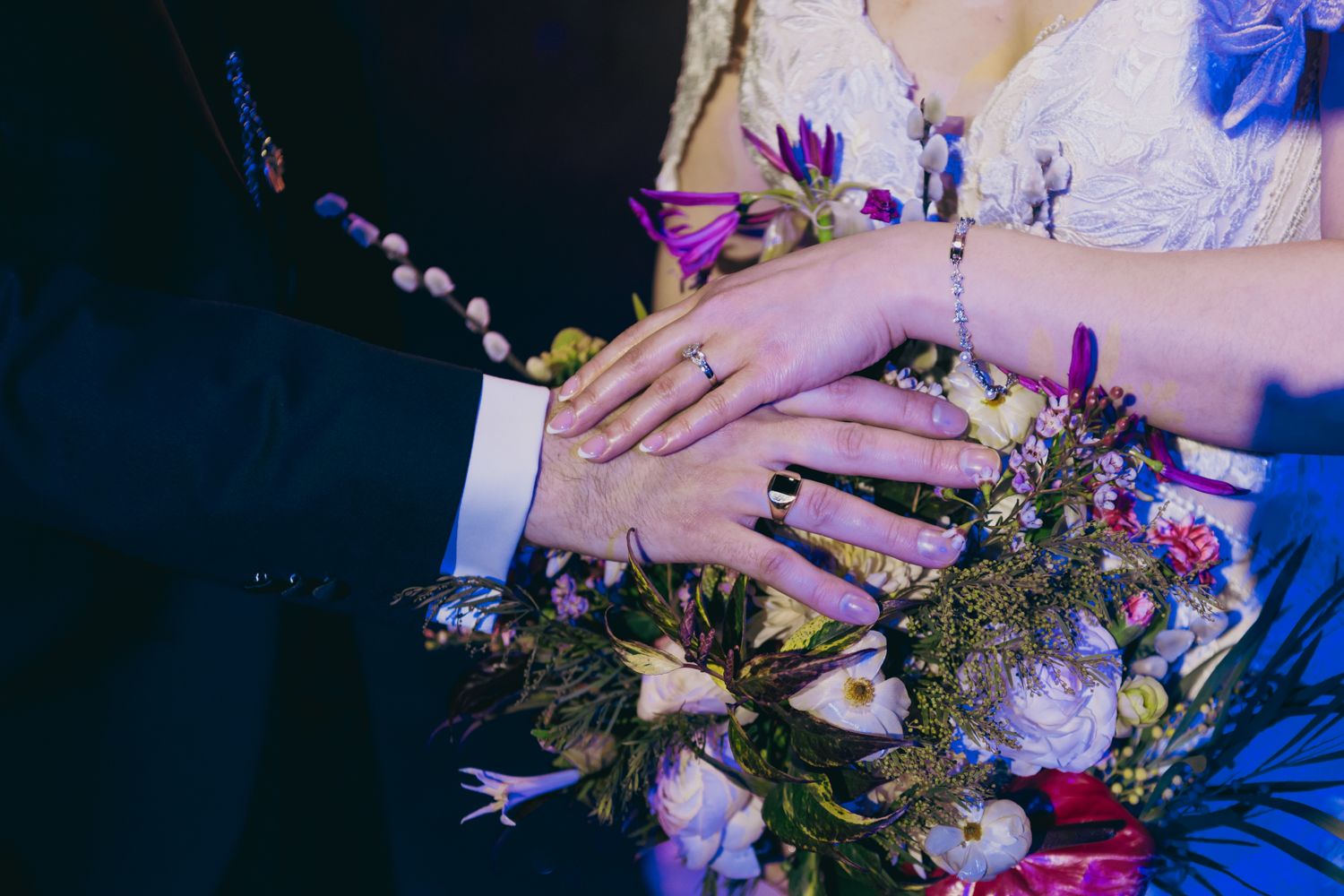 Bride and groom display wedding rings while holding a purple and white floral bouquet together.