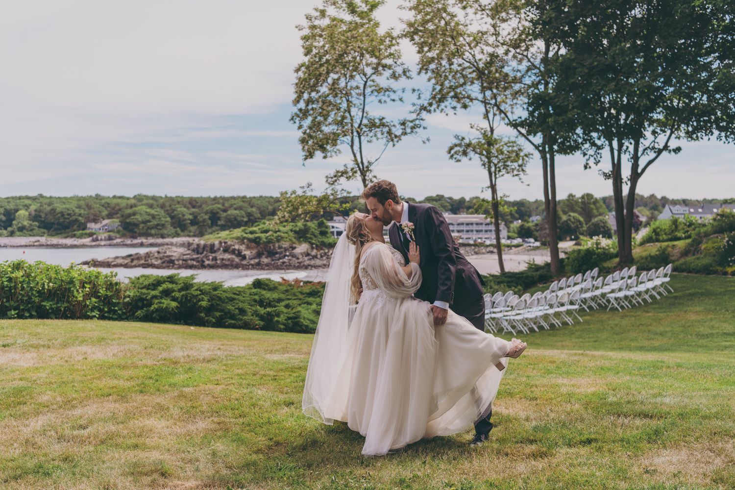 Bride and groom share a romantic kiss on a green lawn with a waterfront view at an outdoor wedding ceremony.