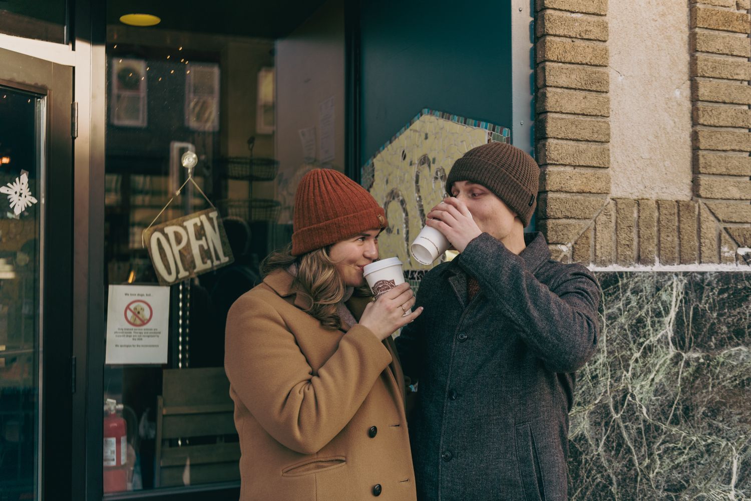 Couple drinking hot beverages outside an open coffee shop in winter coats and knit hats on a cold day.