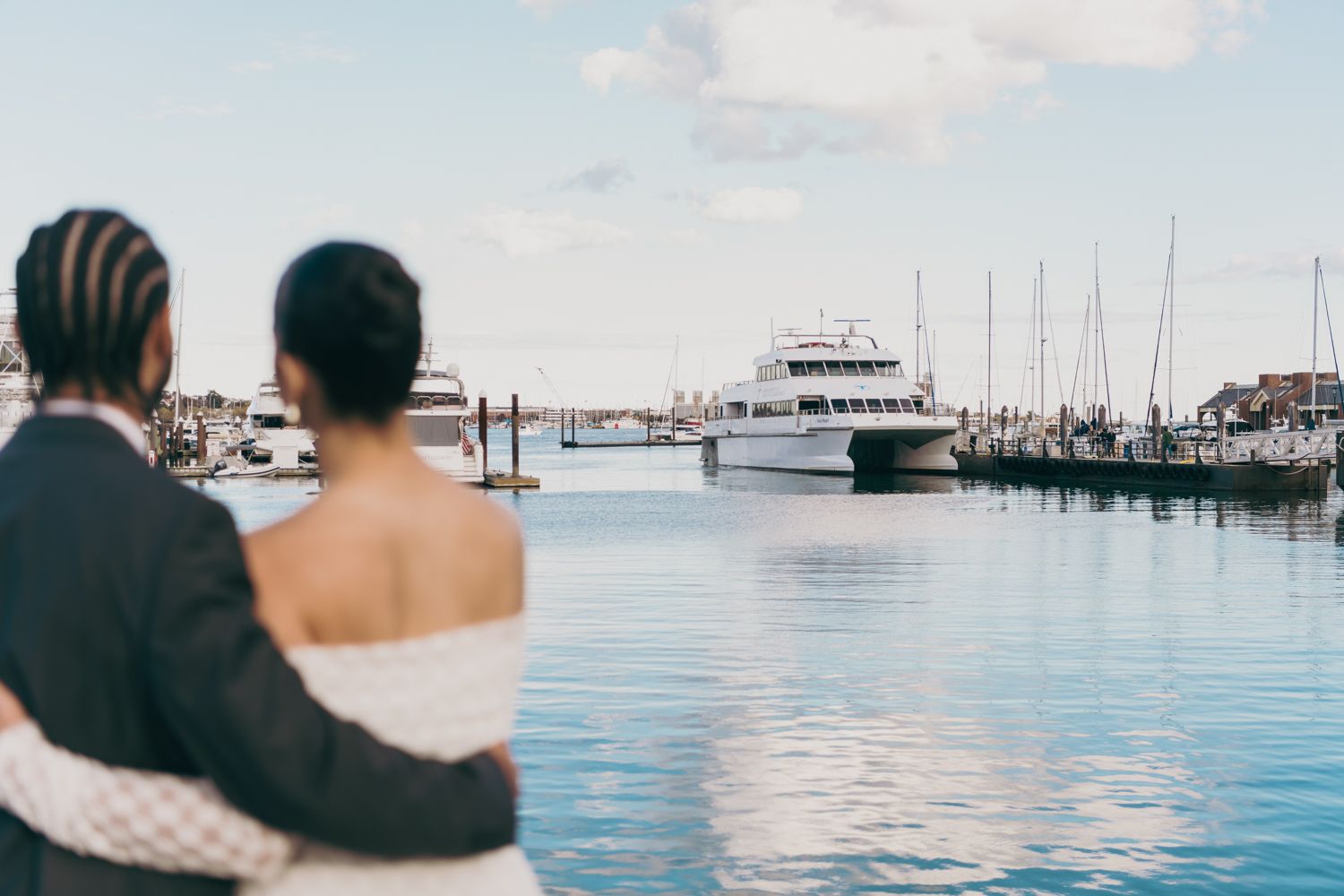 Bride and groom stand together gazing at a yacht docked in a marina on their wedding day.