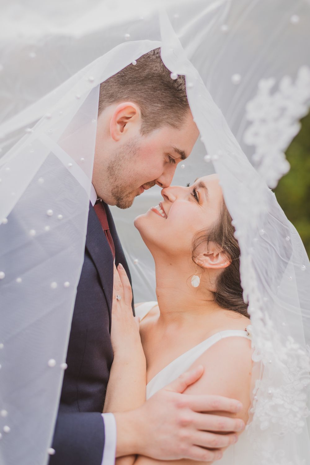 A bride and groom share a romantic moment under a lace wedding veil, nearly kissing and smiling together.
