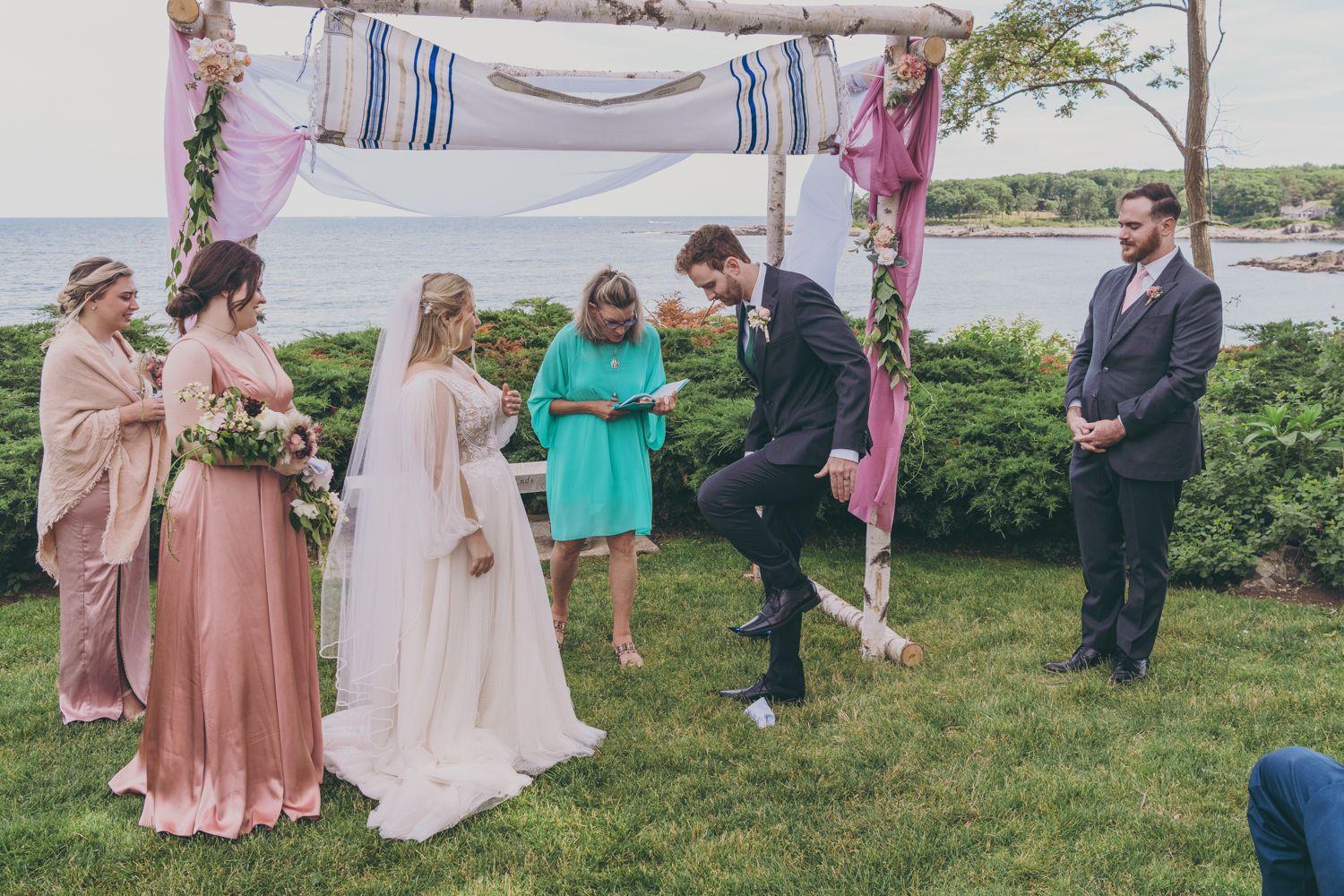 Couple exchanges vows under floral chuppah with bridesmaids and officiant at outdoor waterfront wedding.