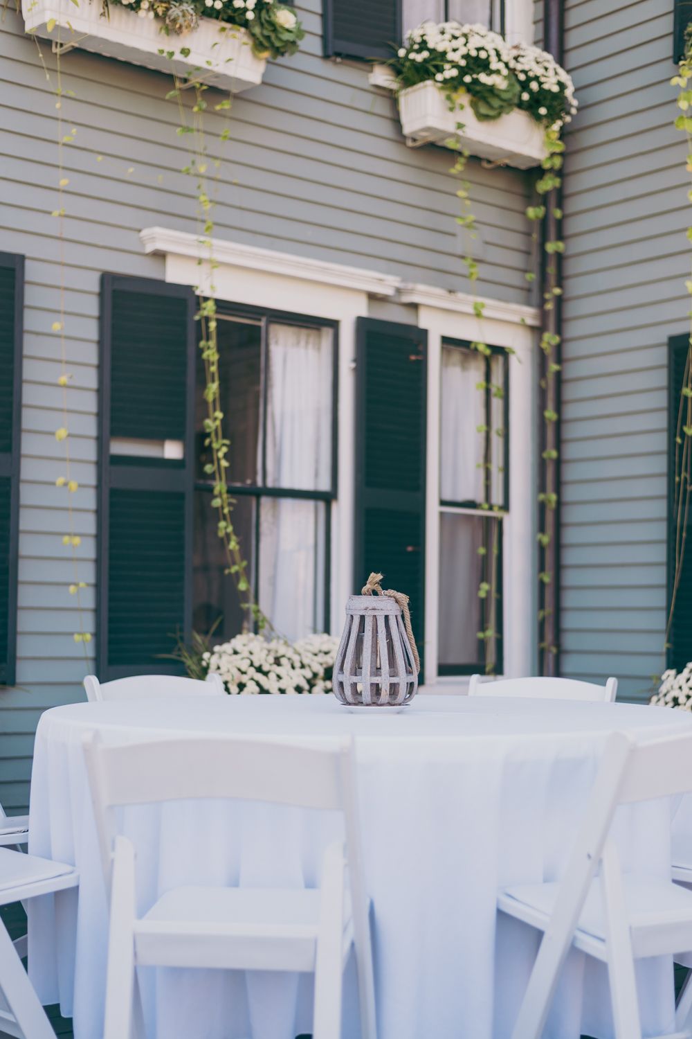 Elegant outdoor wedding reception table with white linen, lantern centerpiece, and floral arrangements against a gray house.