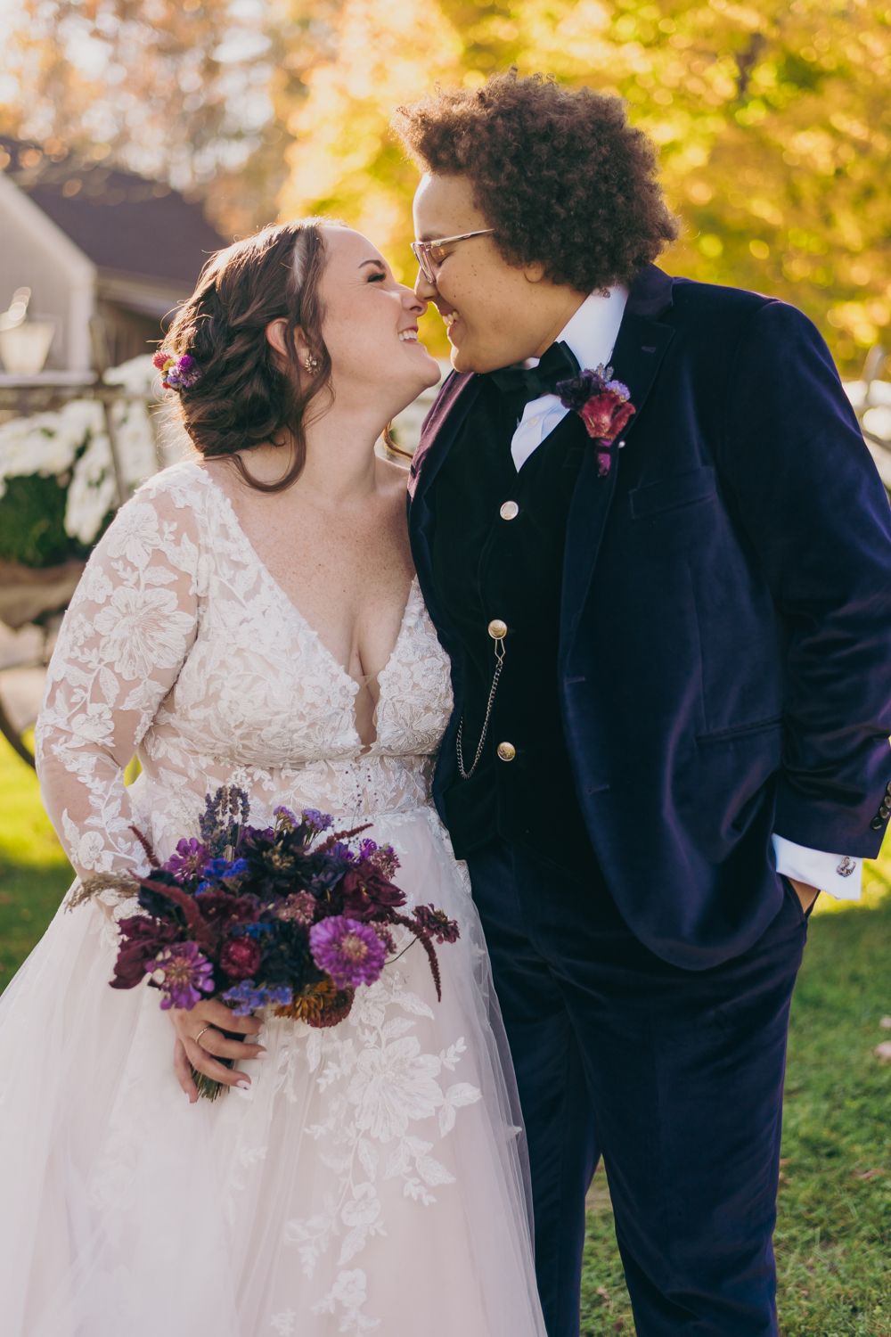 Bride in lace gown holding purple bouquet shares romantic moment with groom in navy suit.