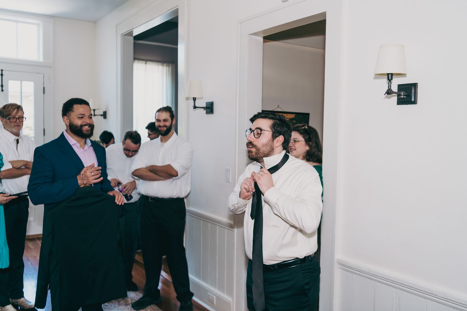 Groom adjusting tie while groomsmen in navy suits laugh and celebrate before the wedding ceremony.