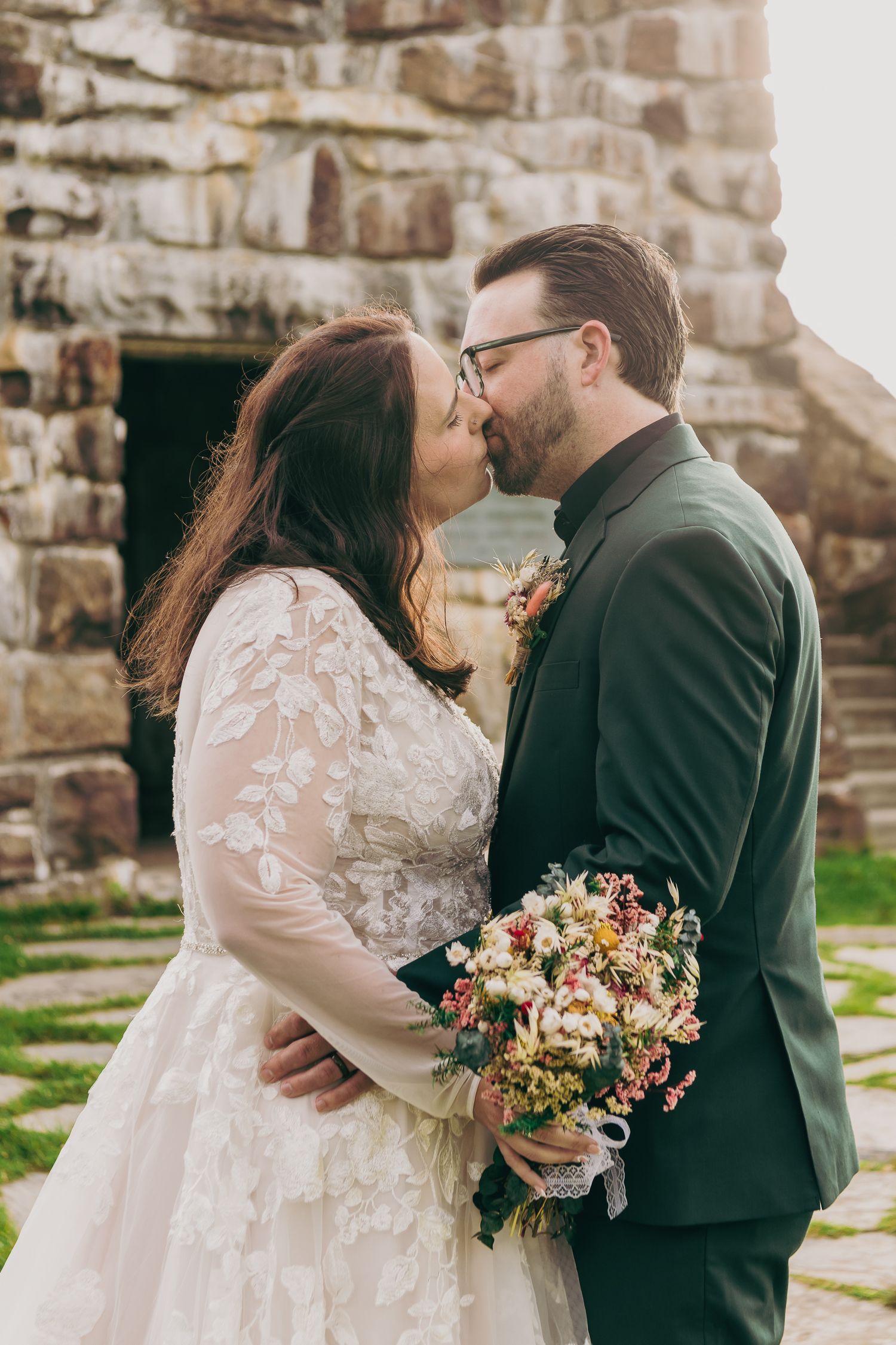 A bride and groom share a romantic kiss outdoors, she holds a colorful bouquet, wearing lace gown, he in dark green suit.