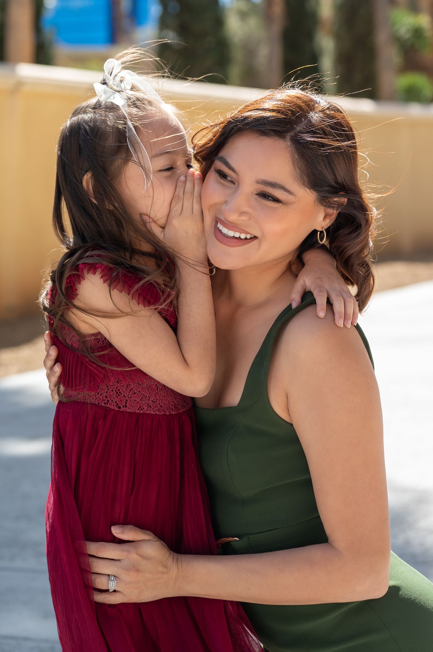 A young girl in a red dress whispers in the ear of a smiling woman in a green dress outdoors.