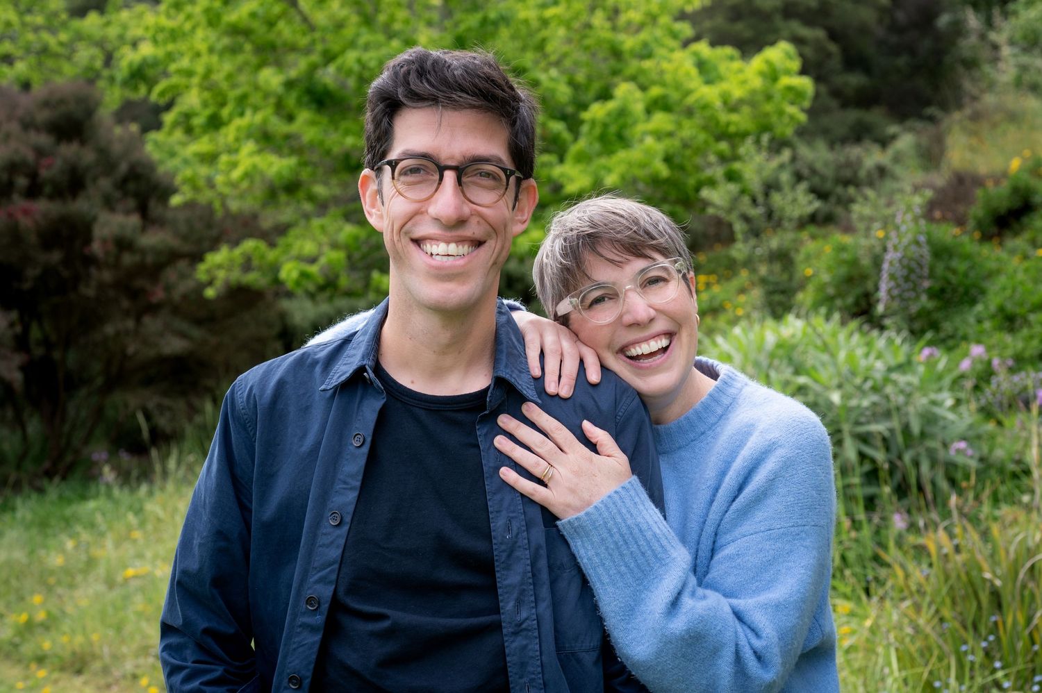 Smiling couple posing together outdoors surrounded by lush green trees.