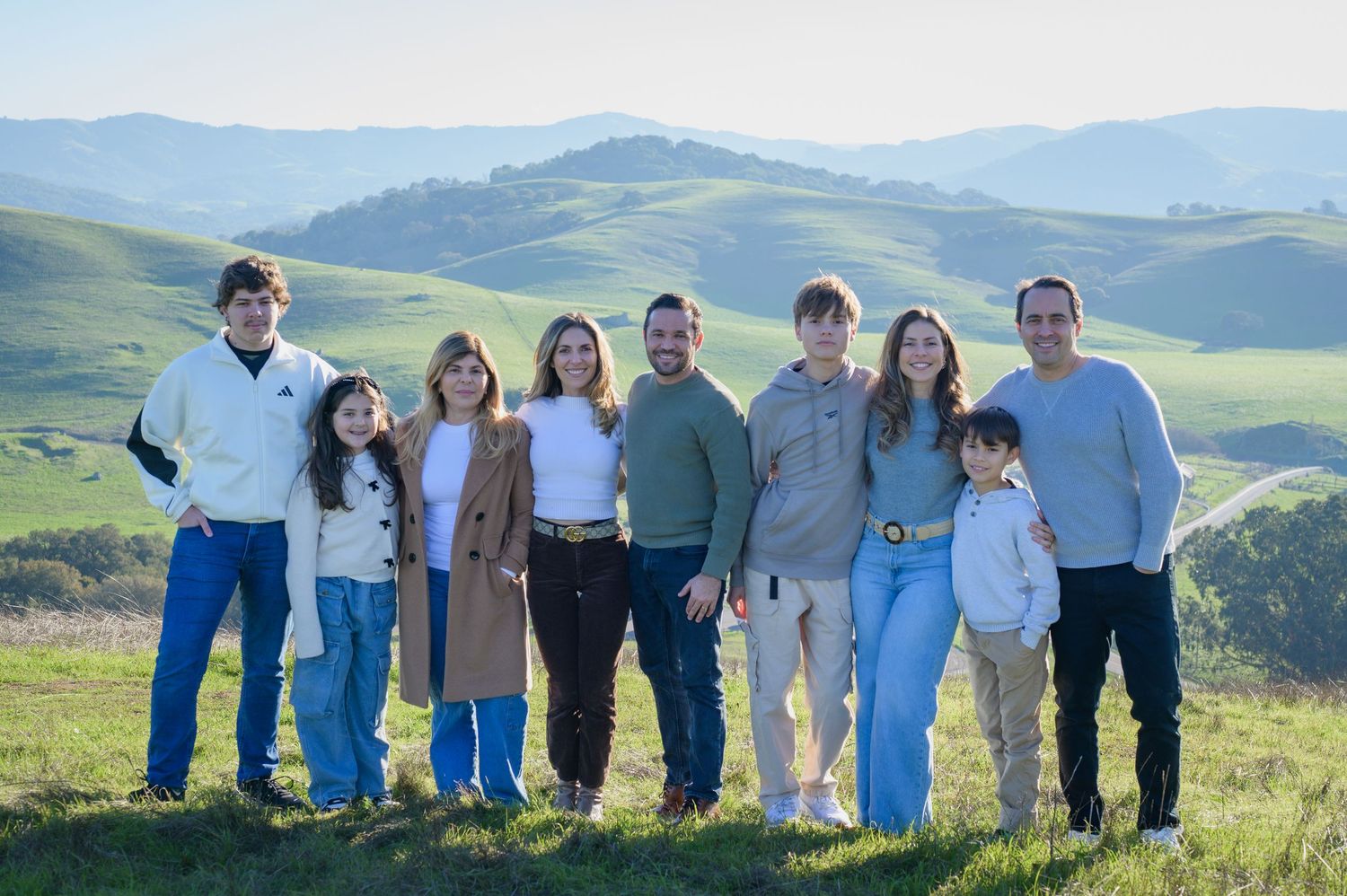 A large family of eight poses together on a lush green hillside with rolling hills and blue sky behind them.