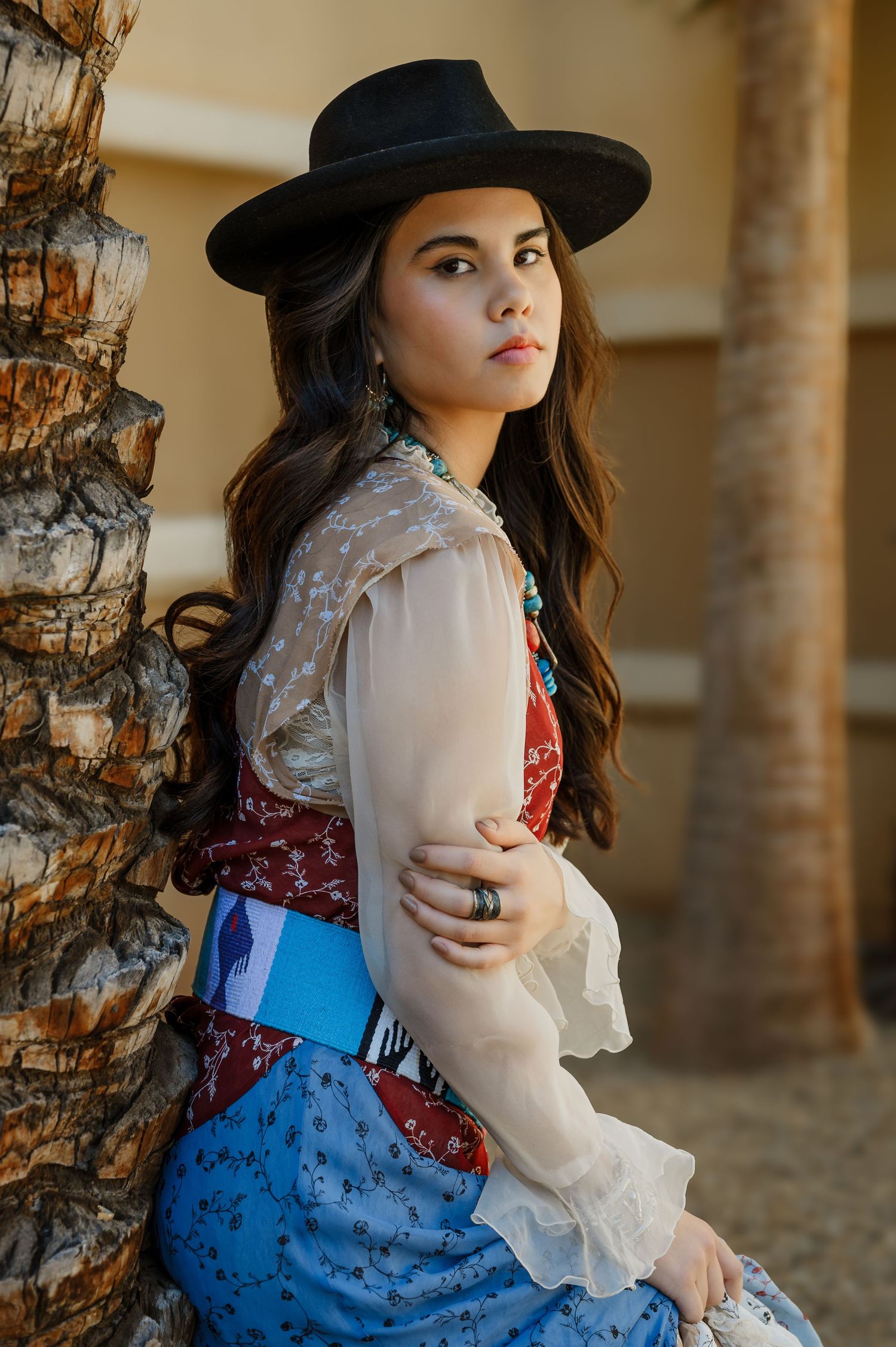 Young woman in black wide-brim hat and boho southwestern outfit poses gracefully beside a palm tree outdoors.