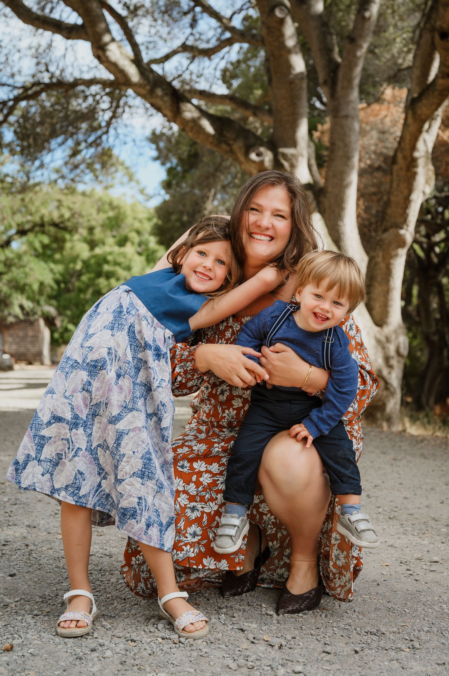 A smiling mother crouches outdoors hugging her two young children, a girl in a blue dress and a boy in a navy top.
