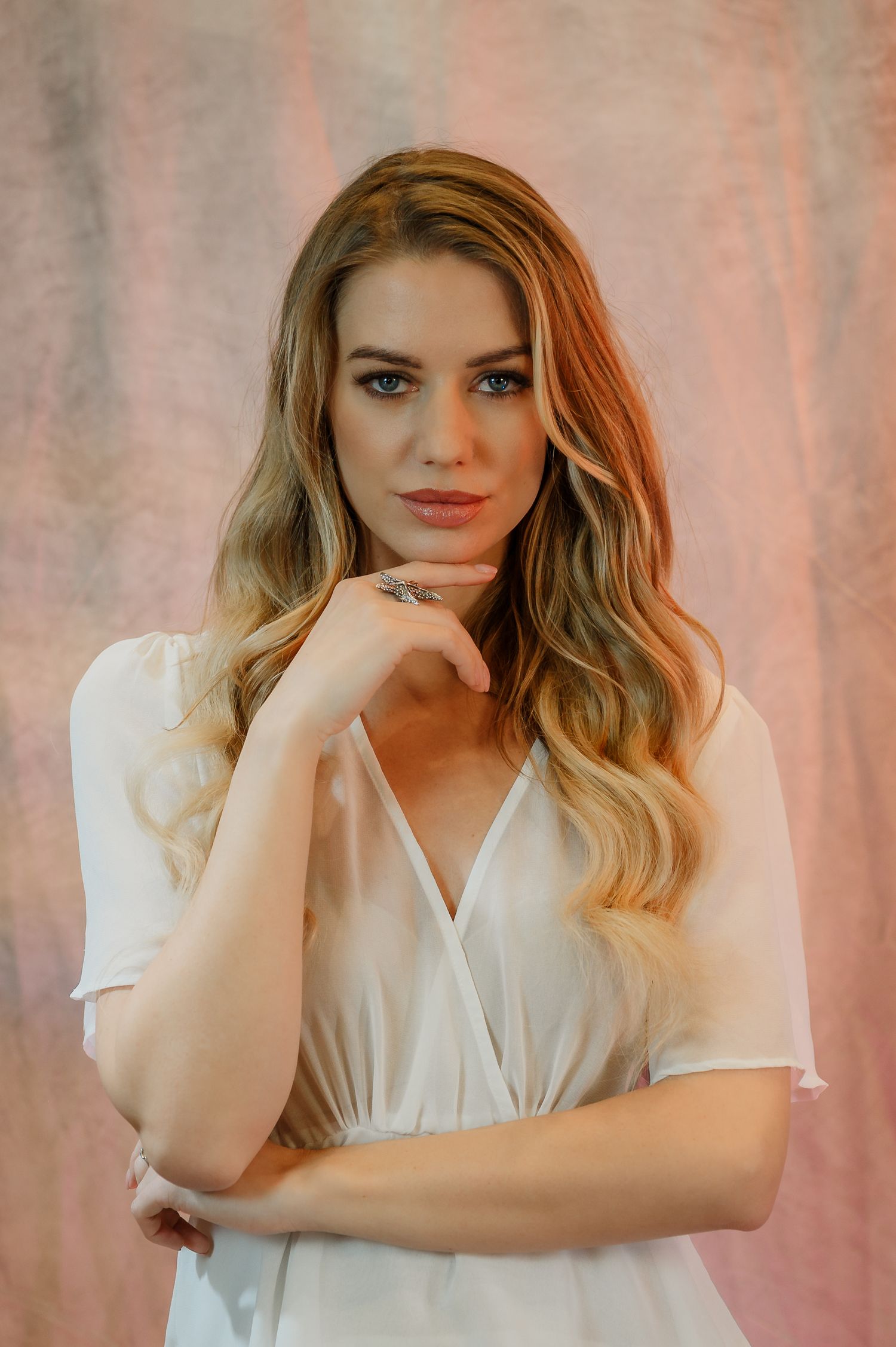 A young blonde woman in a white wrap dress poses thoughtfully with hand on chin against a soft pink backdrop.