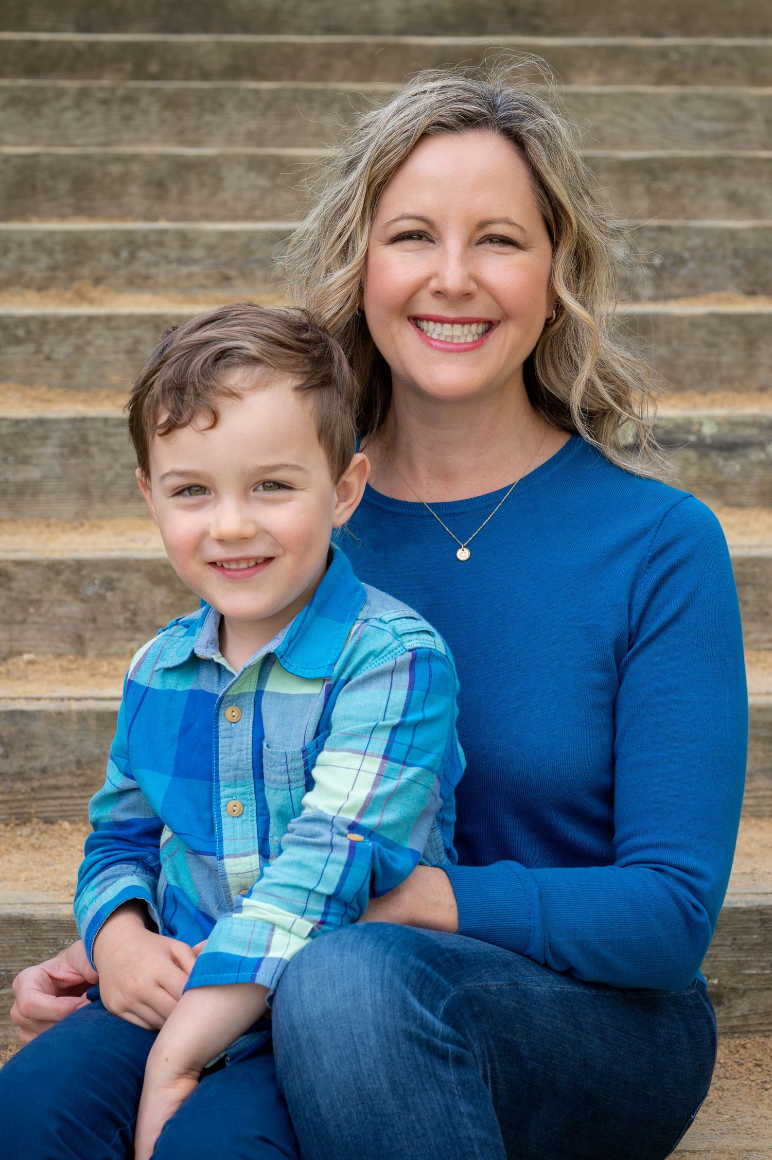 A smiling woman in a blue sweater sits on outdoor steps with a young boy in a plaid shirt, both looking happy.