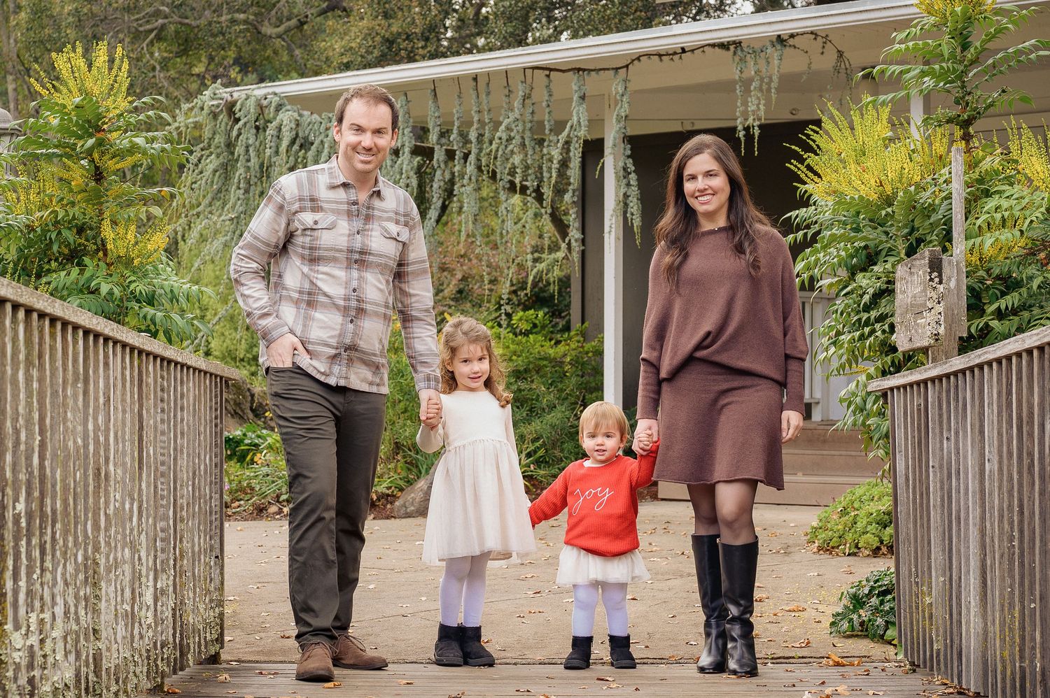 A happy family of four poses on a garden path, with two young children holding hands between their smiling parents.