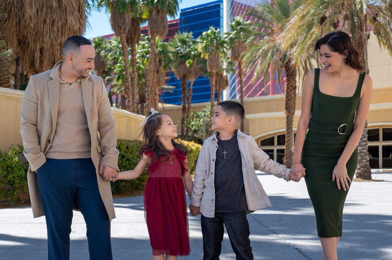 Happy family of four holding hands outdoors near palm trees and colorful buildings on a sunny day.