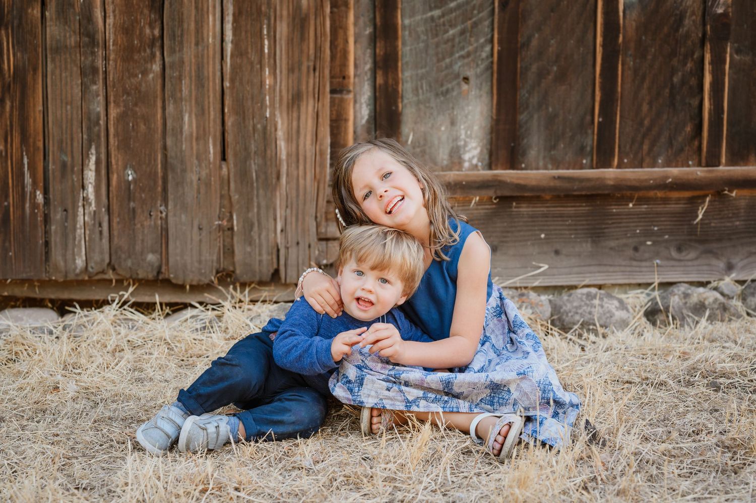 Two young siblings hugging and smiling while sitting on hay in front of a rustic wooden barn wall.