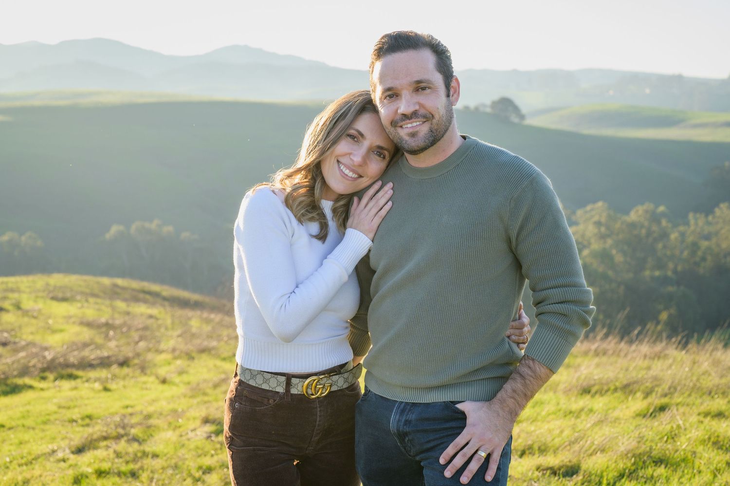 Couple embracing outdoors on a green hillside, woman in white top leaning on man in olive sweater.