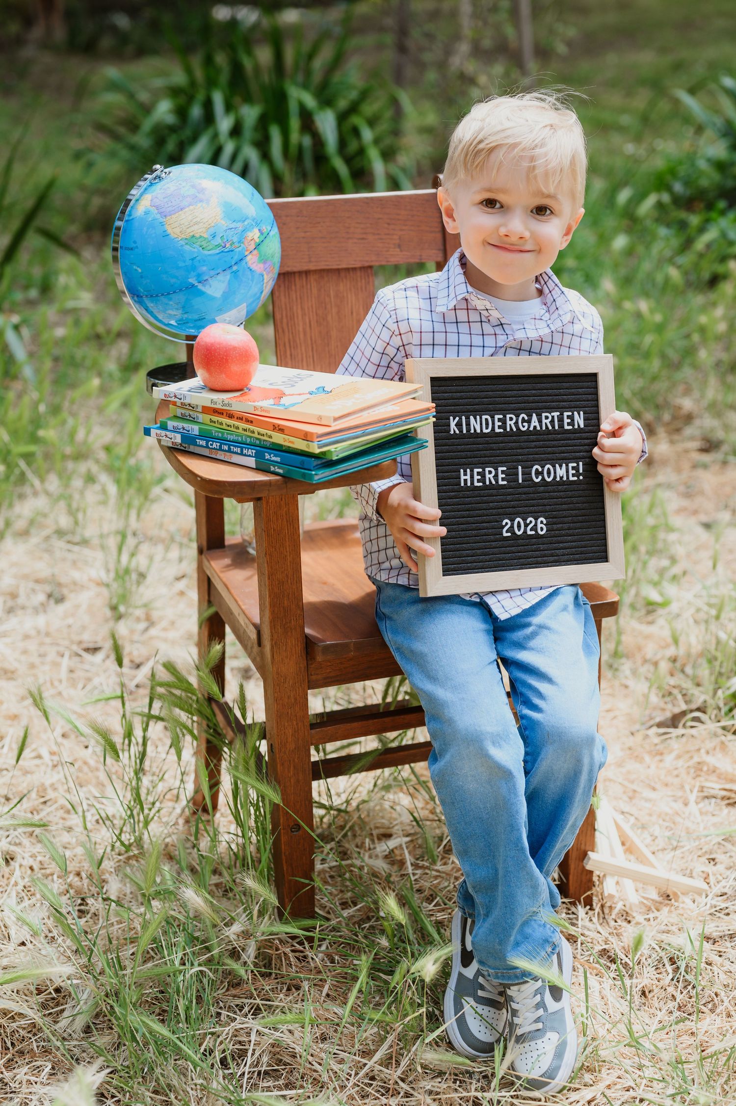 Smiling blond boy holding a letter board sign reading Kindergarten Here I Come 2026 seated on a wooden chair outdoors.