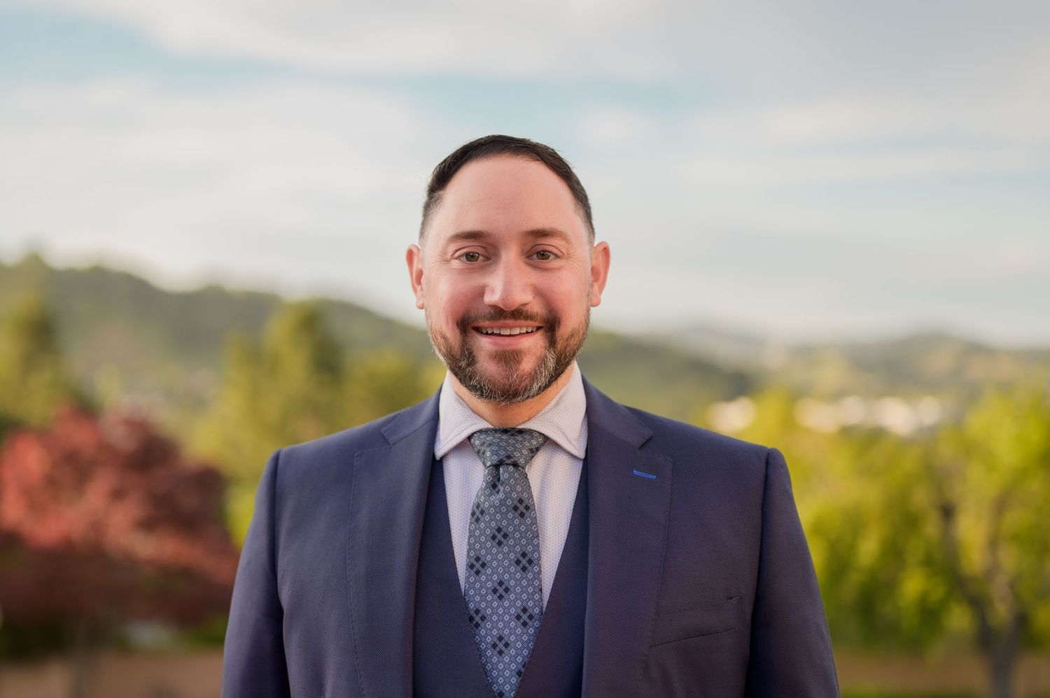 Professional man in navy blue suit and tie smiling outdoors with green hills in background.