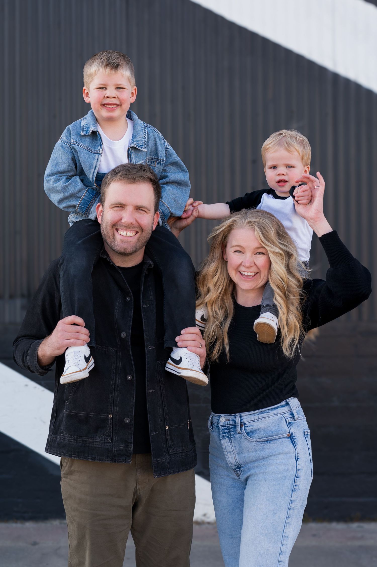 Happy family of four posing outdoors, parents carrying two young blonde boys on their shoulders, smiling.