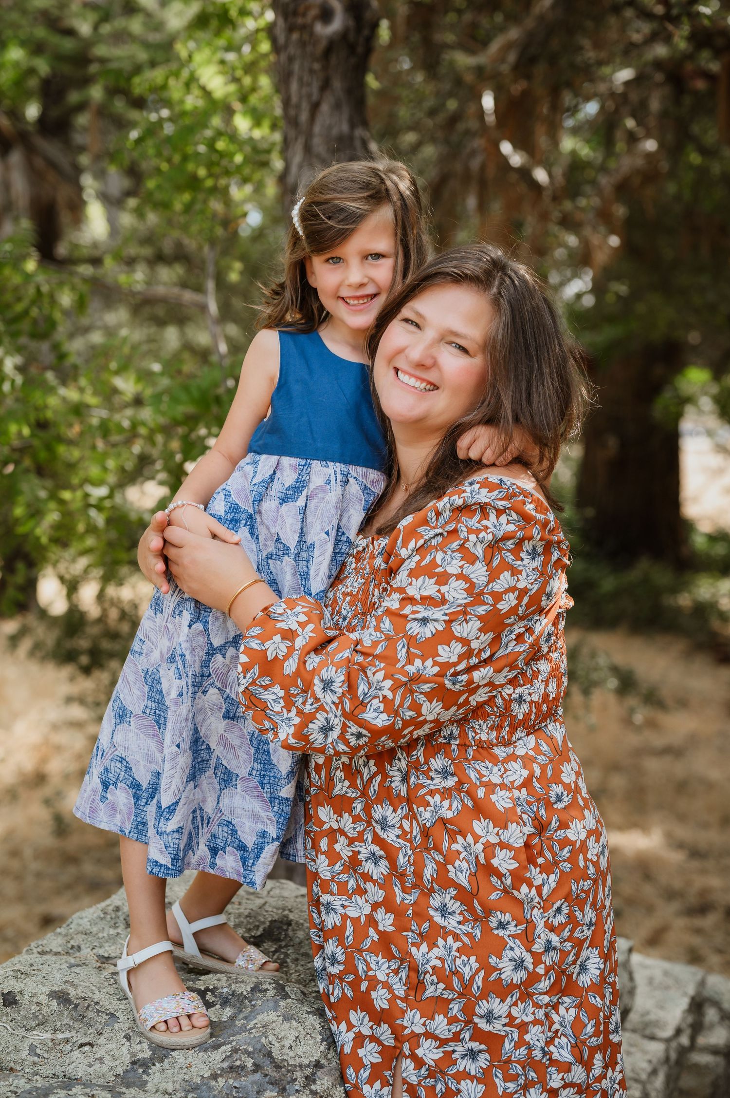 A smiling woman in a floral orange dress holds a young girl in a blue dress outdoors among green trees.
