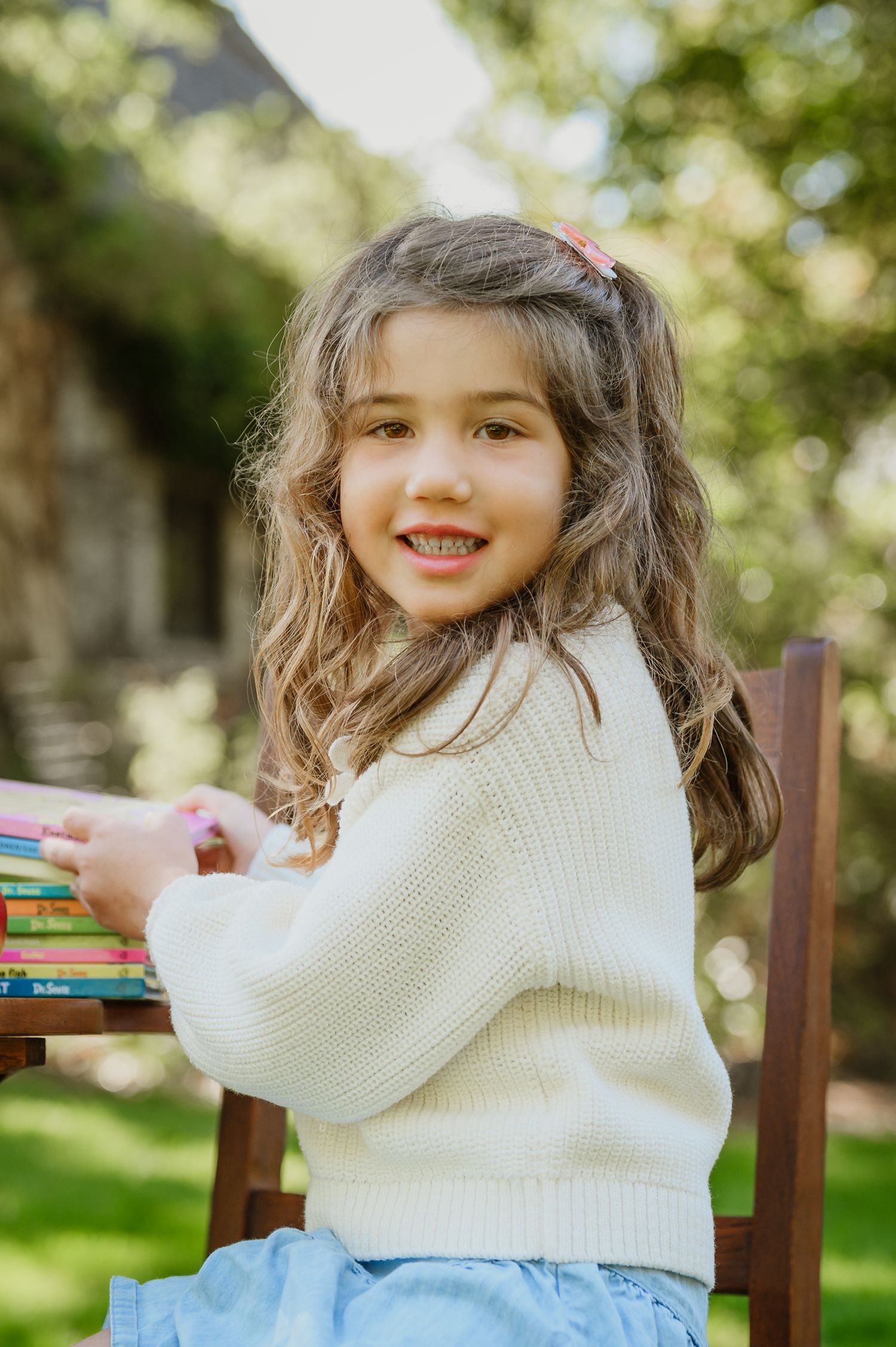 Smiling young girl with curly hair in cream sweater sitting outdoors with colorful books on a wooden chair.