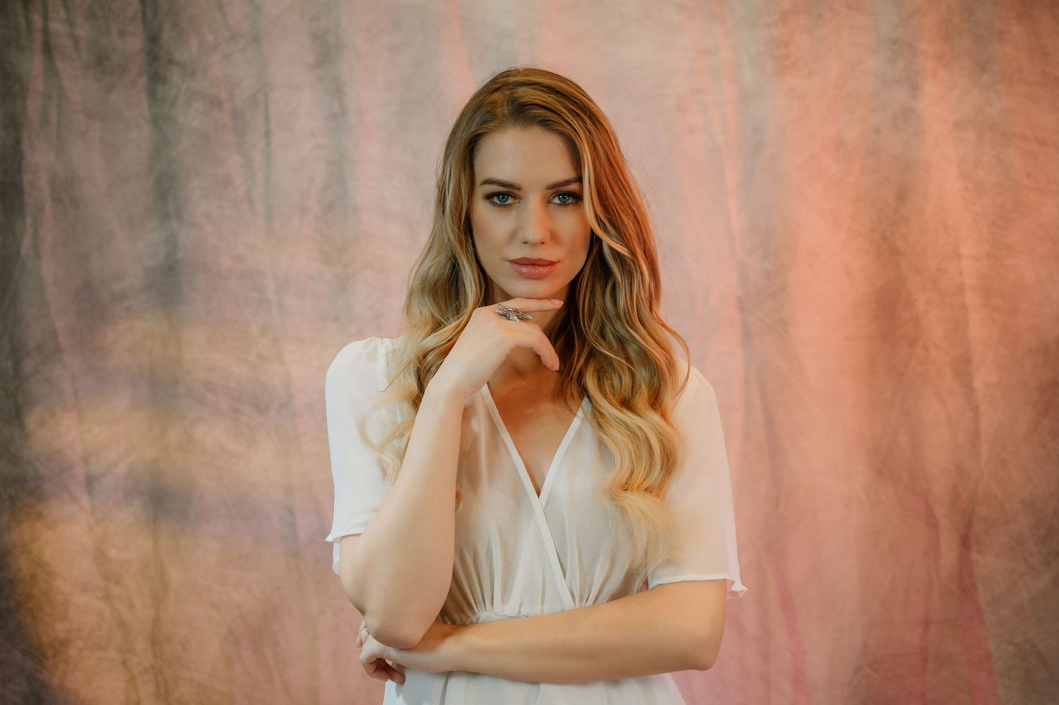 Young woman with blonde wavy hair poses confidently in white wrap top against soft pink textured backdrop.