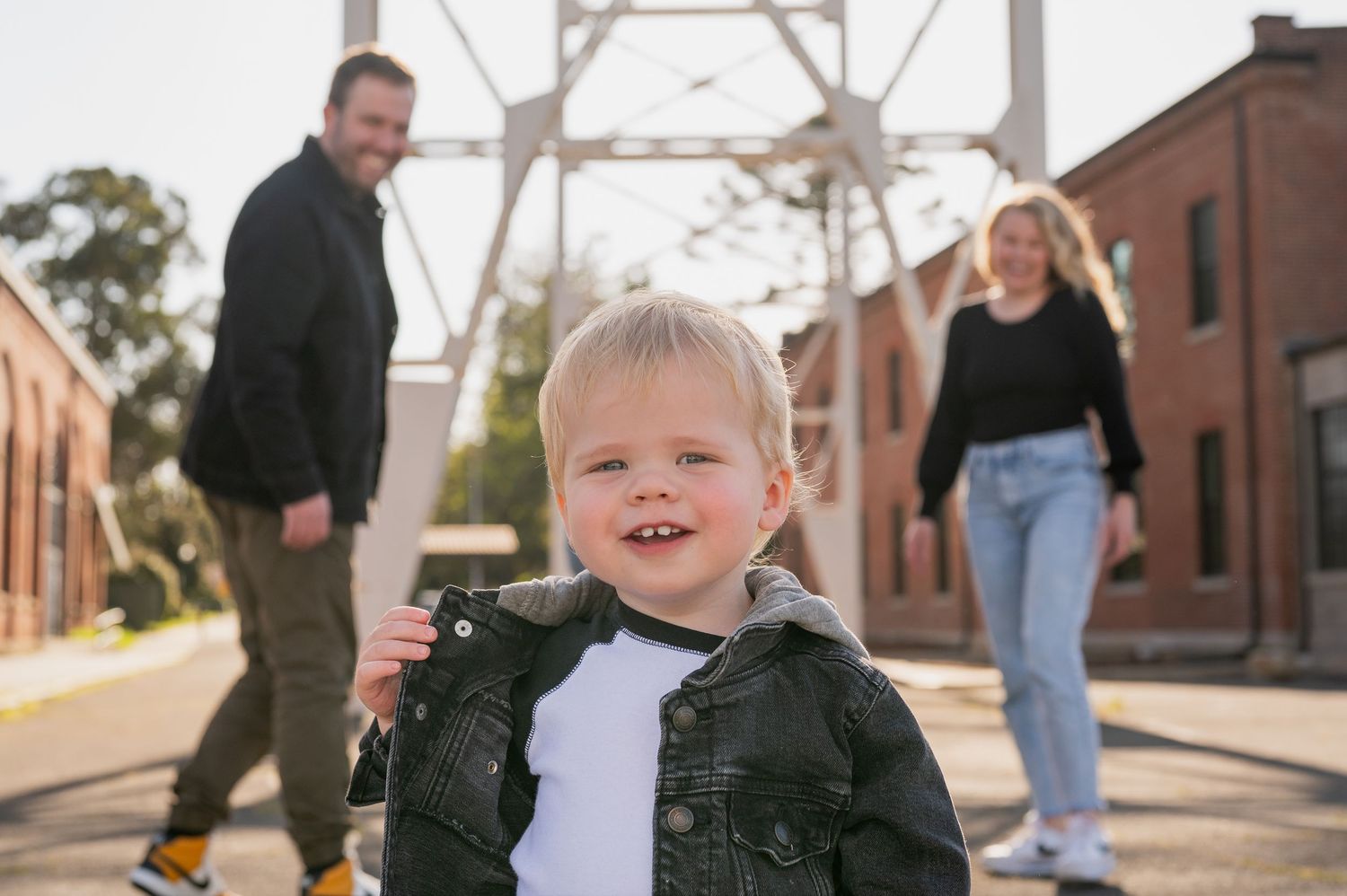 Smiling toddler in denim jacket with parents standing behind outdoors.