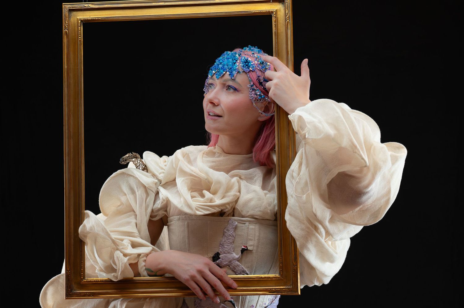 Woman in white ruffled costume holds golden picture frame, wearing blue floral headscarf against black background.