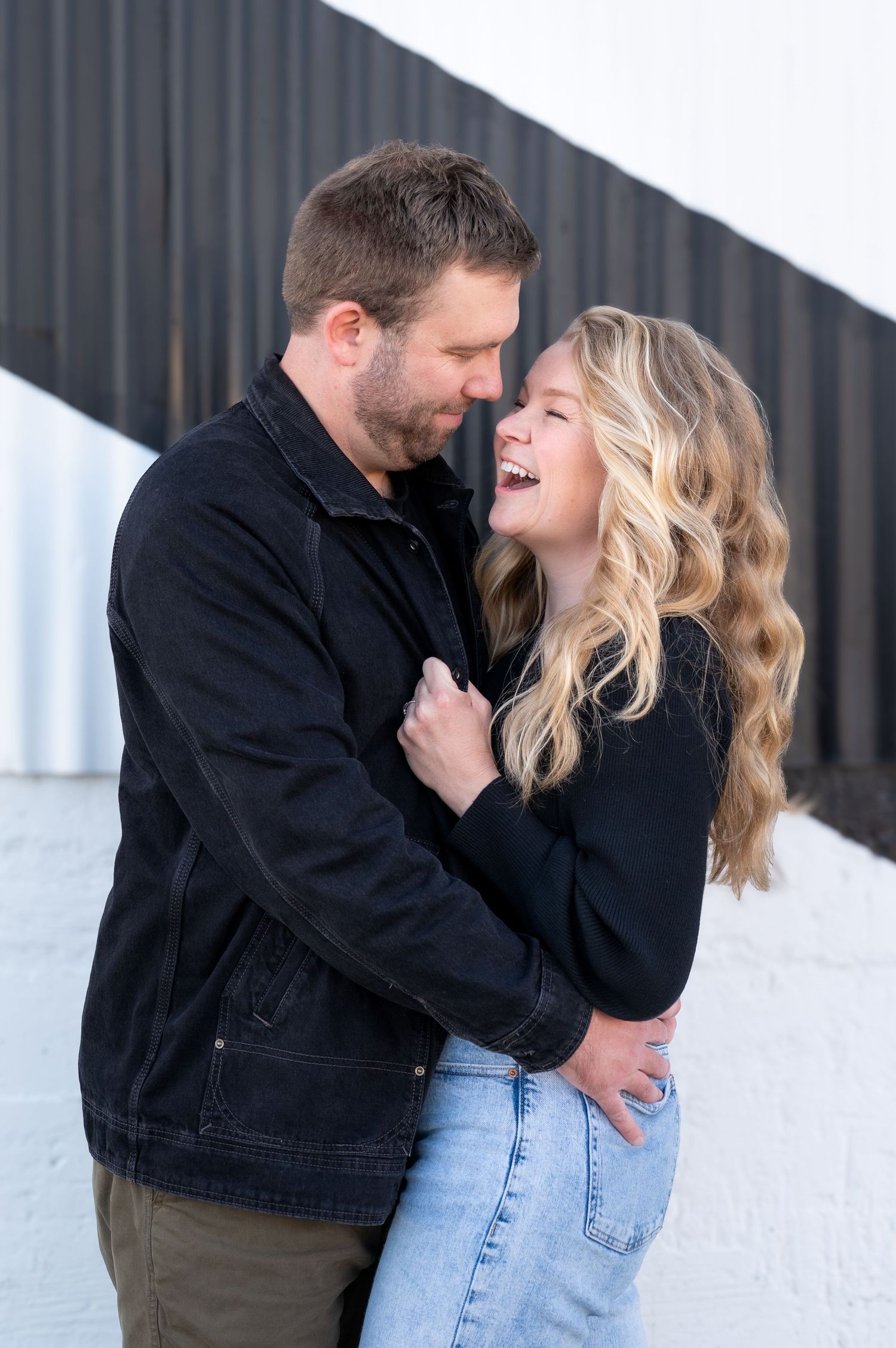Laughing couple embracing outdoors against white and black wall background.