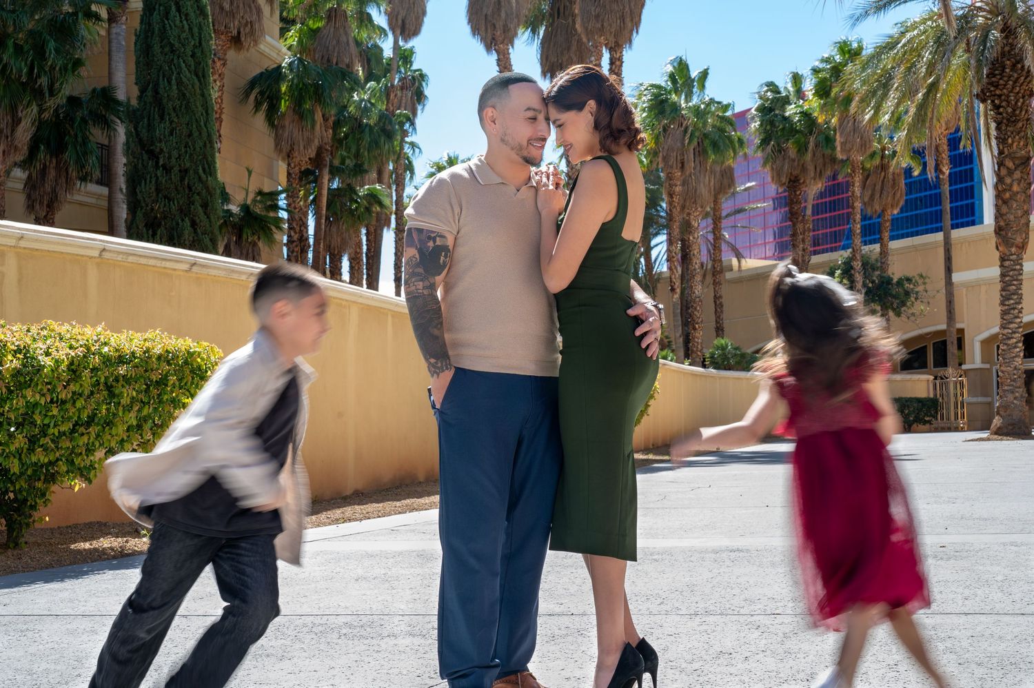 Couple embracing outdoors near palm trees while two children run past in motion blur, Las Vegas backdrop.