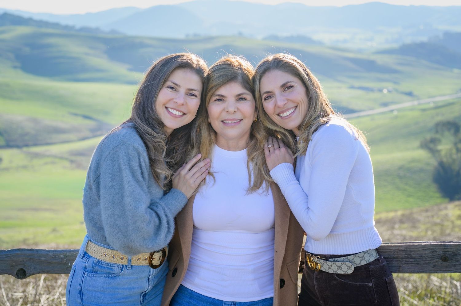 Three smiling women posing together with green rolling hills behind them.