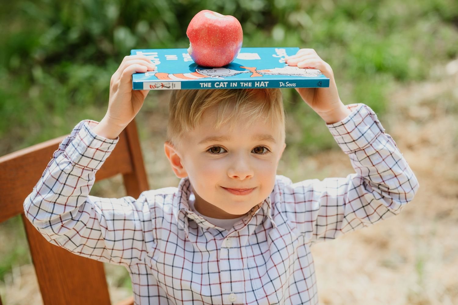 Young boy balancing The Cat in the Hat book and red apple on his head outdoors.