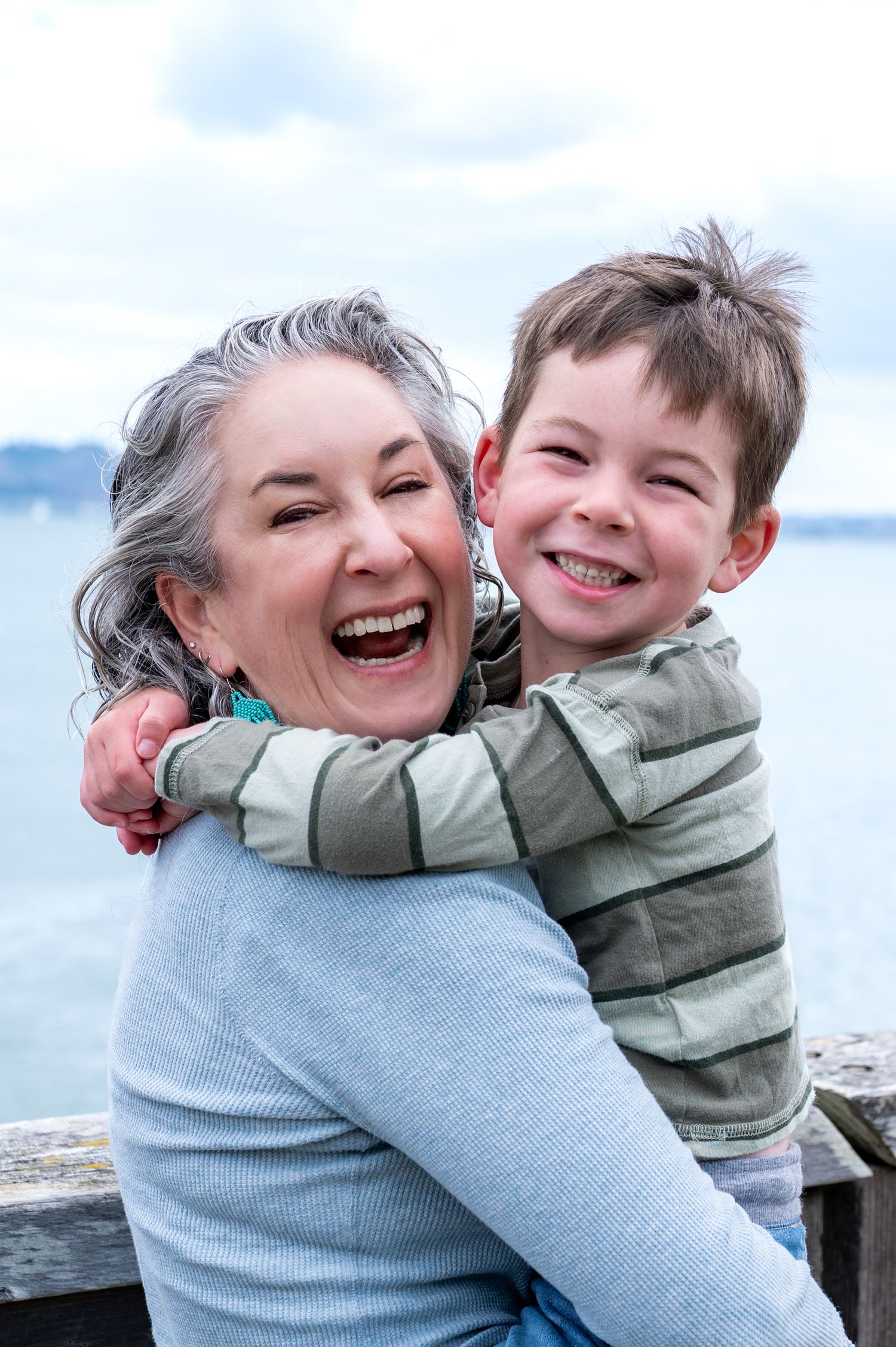 Grandmother and young boy laughing together on a waterfront dock, sharing a joyful hug outdoors.