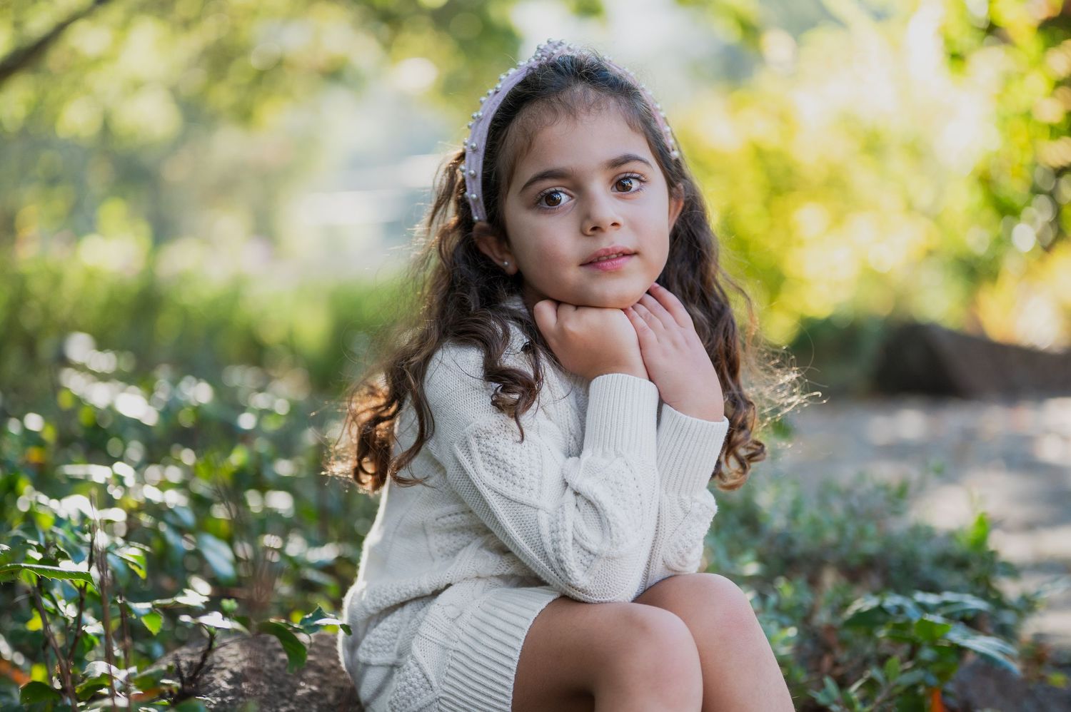 Young girl with curly hair sitting outdoors in a white sweater.