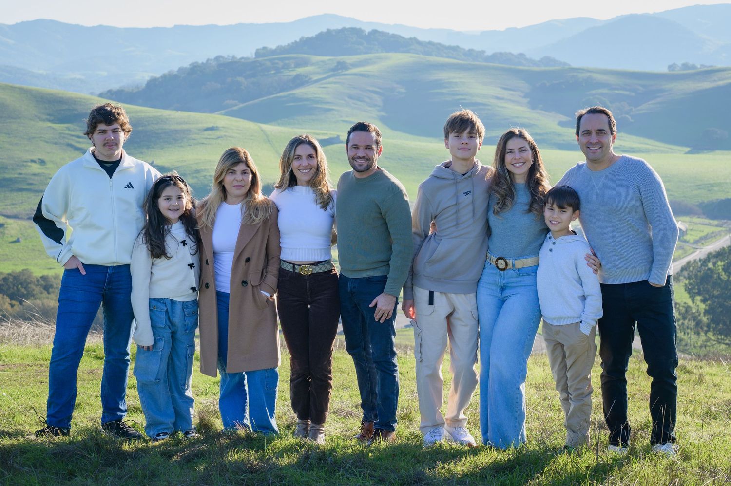 A large family group poses together smiling on a green hillside with rolling valleys in the background.