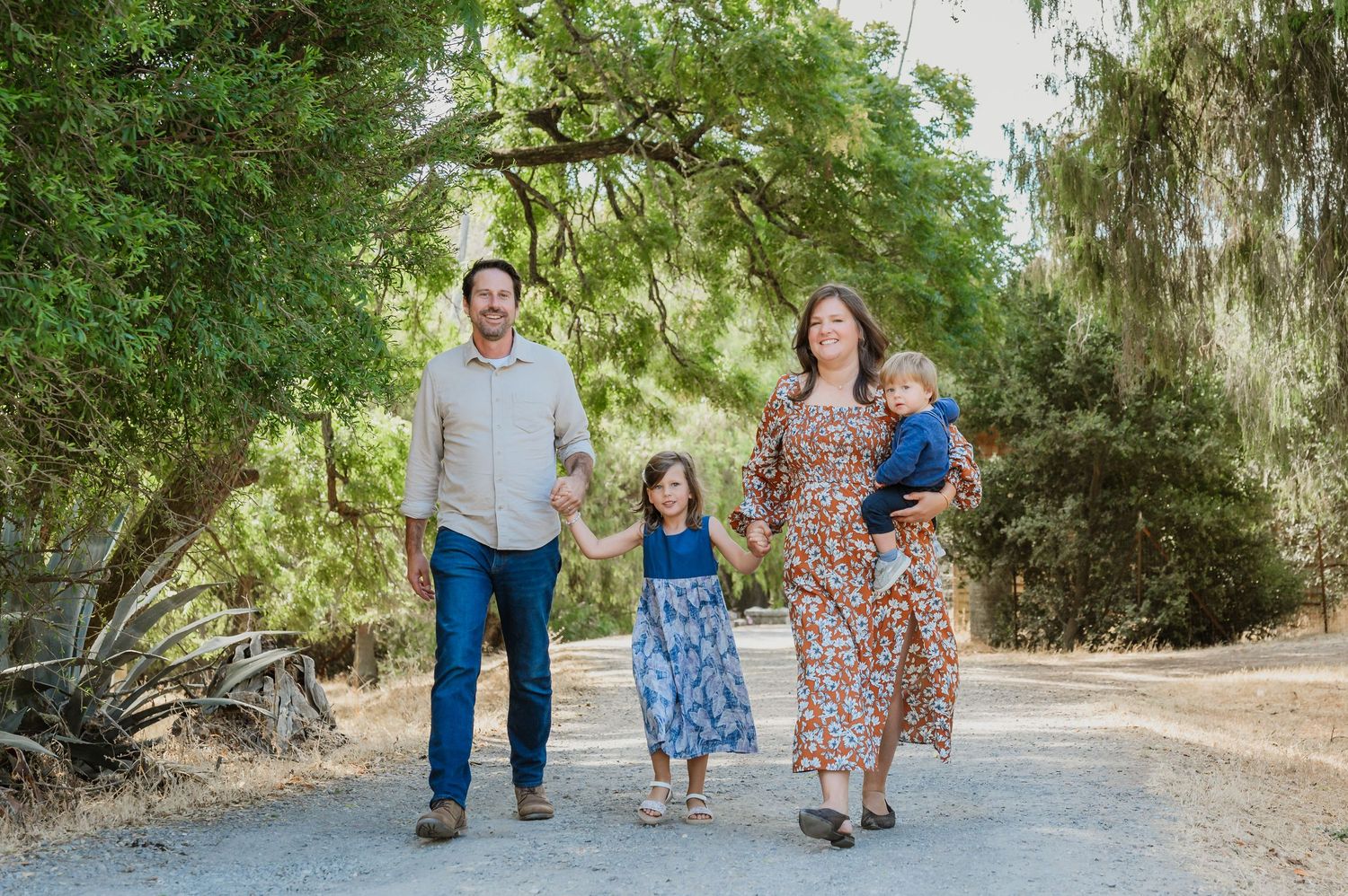 Family of four walks on a sunny dirt path surrounded by lush green trees, parents holding hands with young children.