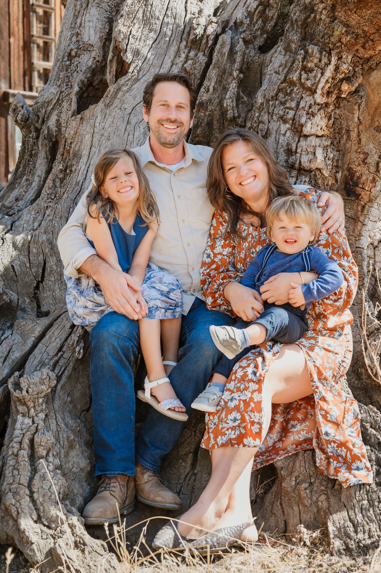 Happy family of four posing together against a large tree trunk outdoors in a warm, natural setting.