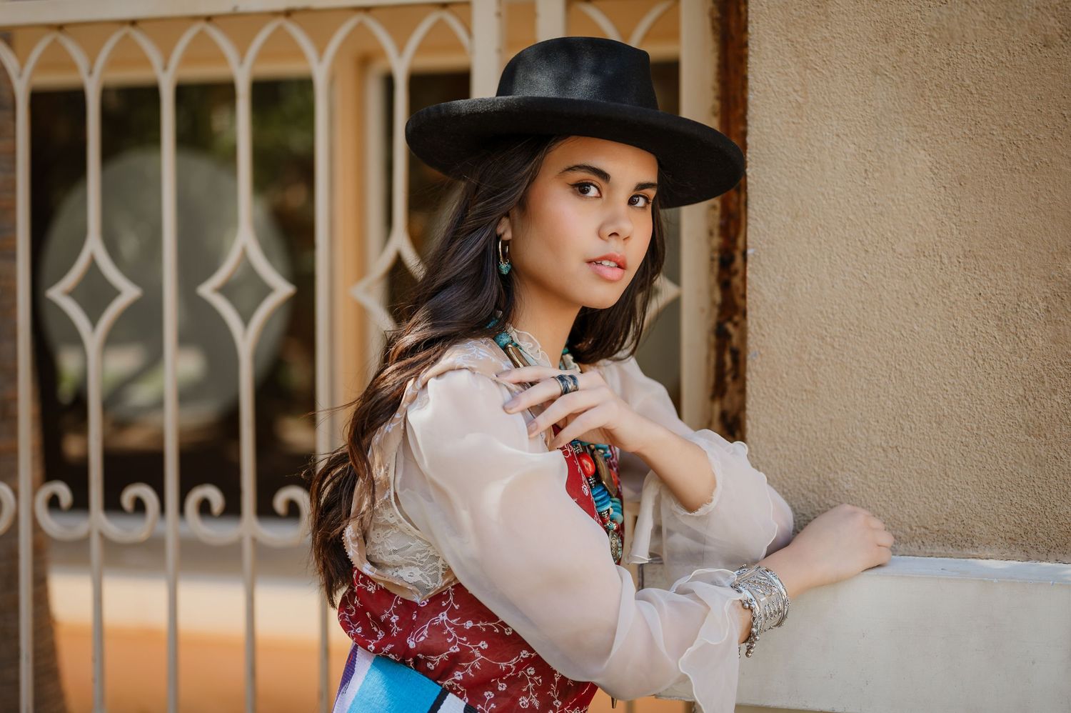 Young woman in black wide-brim hat and boho outfit leaning against wall outdoors.