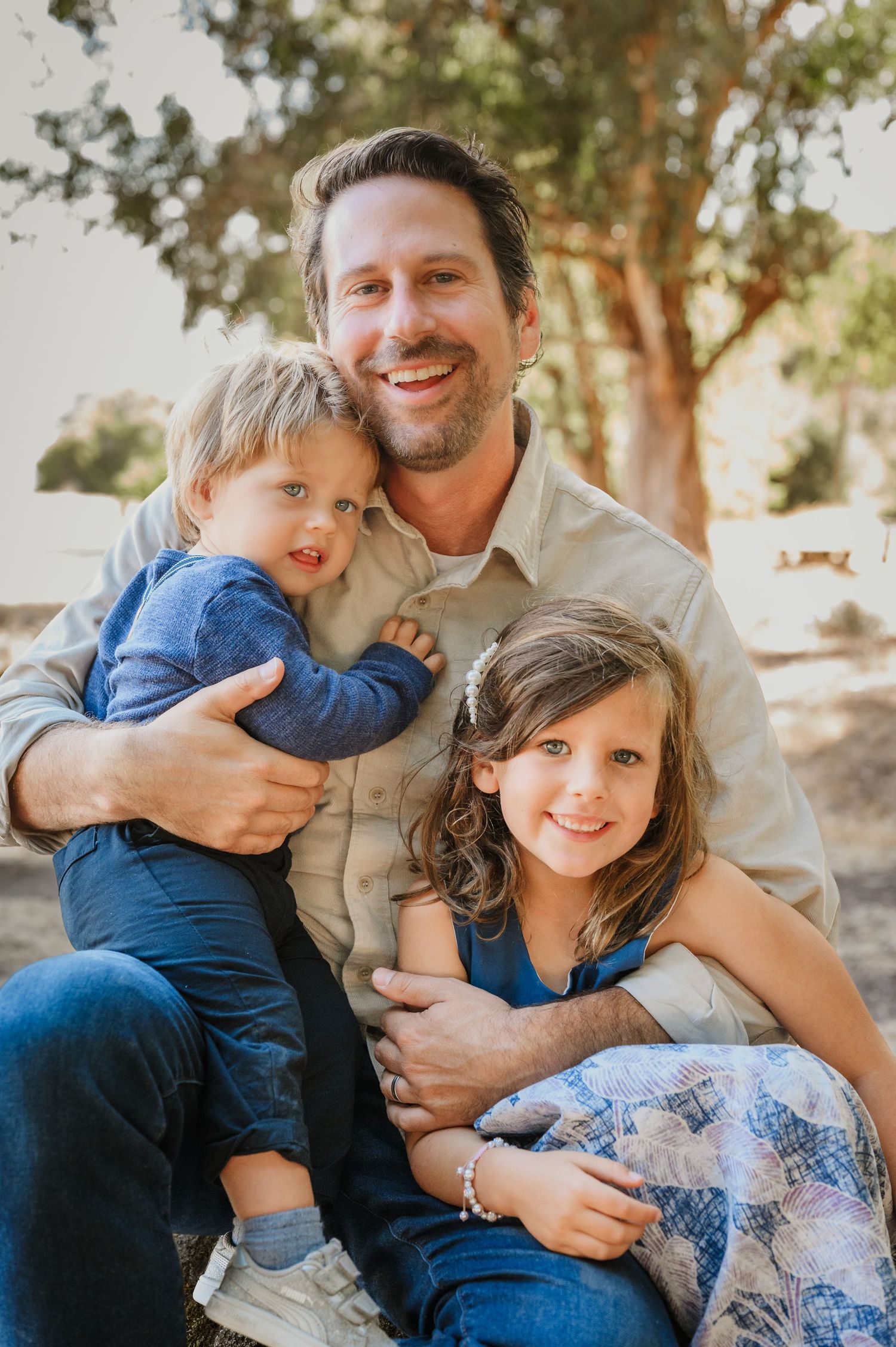 A smiling man sits outdoors holding two young children, a toddler boy and a girl, in a warm family portrait.