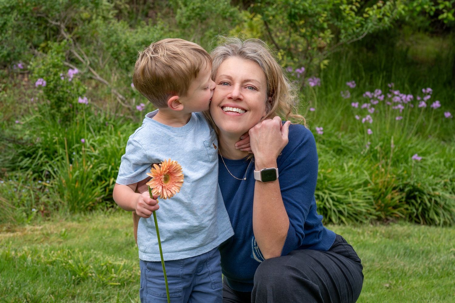 Young boy kissing smiling woman on cheek while holding orange gerbera daisy in lush green garden.