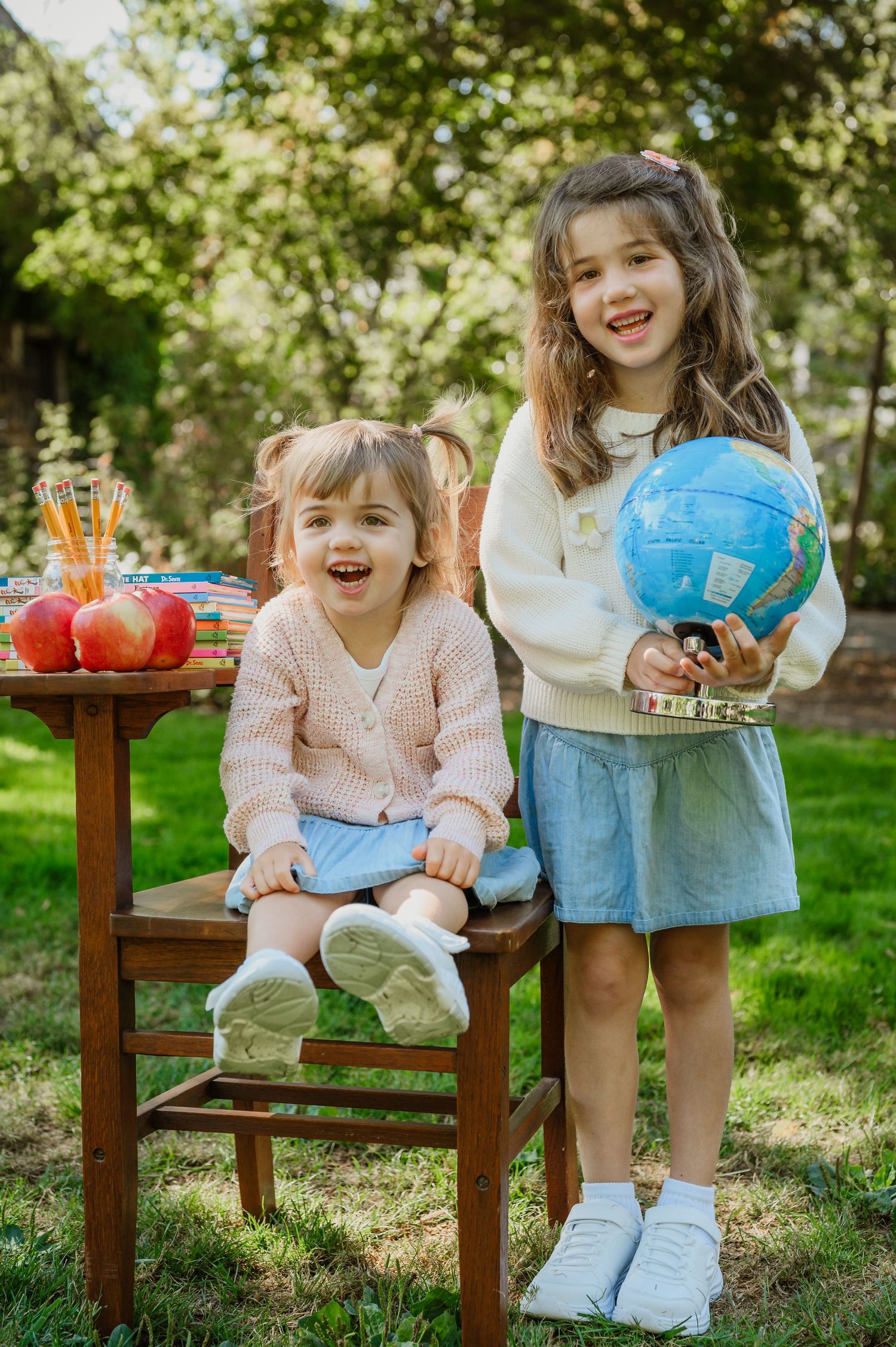 Two young girls smile outdoors, one holding a globe, beside a wooden desk with apples and pencils, back-to-school theme.