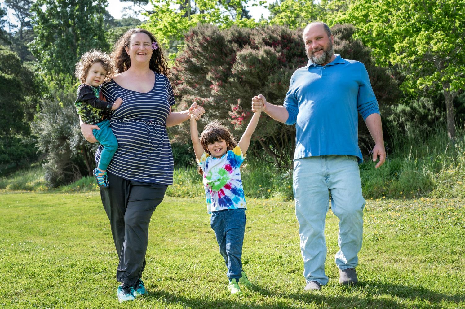 Family of four walking hand-in-hand on green grass in a sunny park, parents carrying and guiding young children.