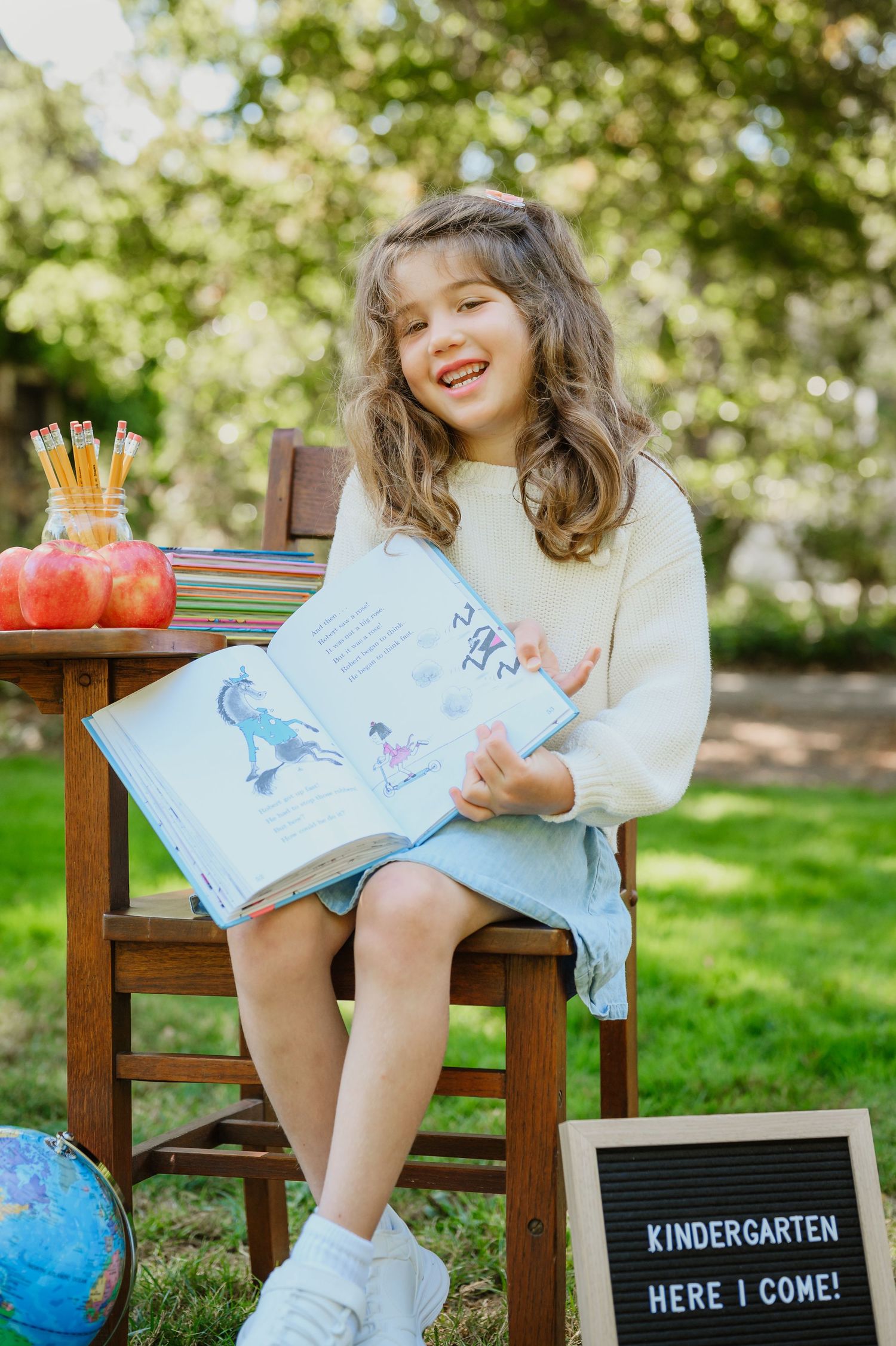 Smiling girl holding an open book outdoors, sitting beside a Kindergarten Here I Come letter board sign.
